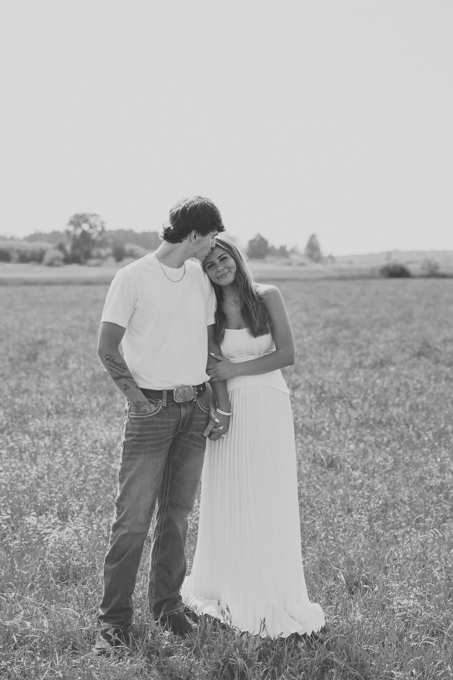 Black and white photo of a romantic couple embracing in an open field, woman in white dress leaning on mans shoulder.