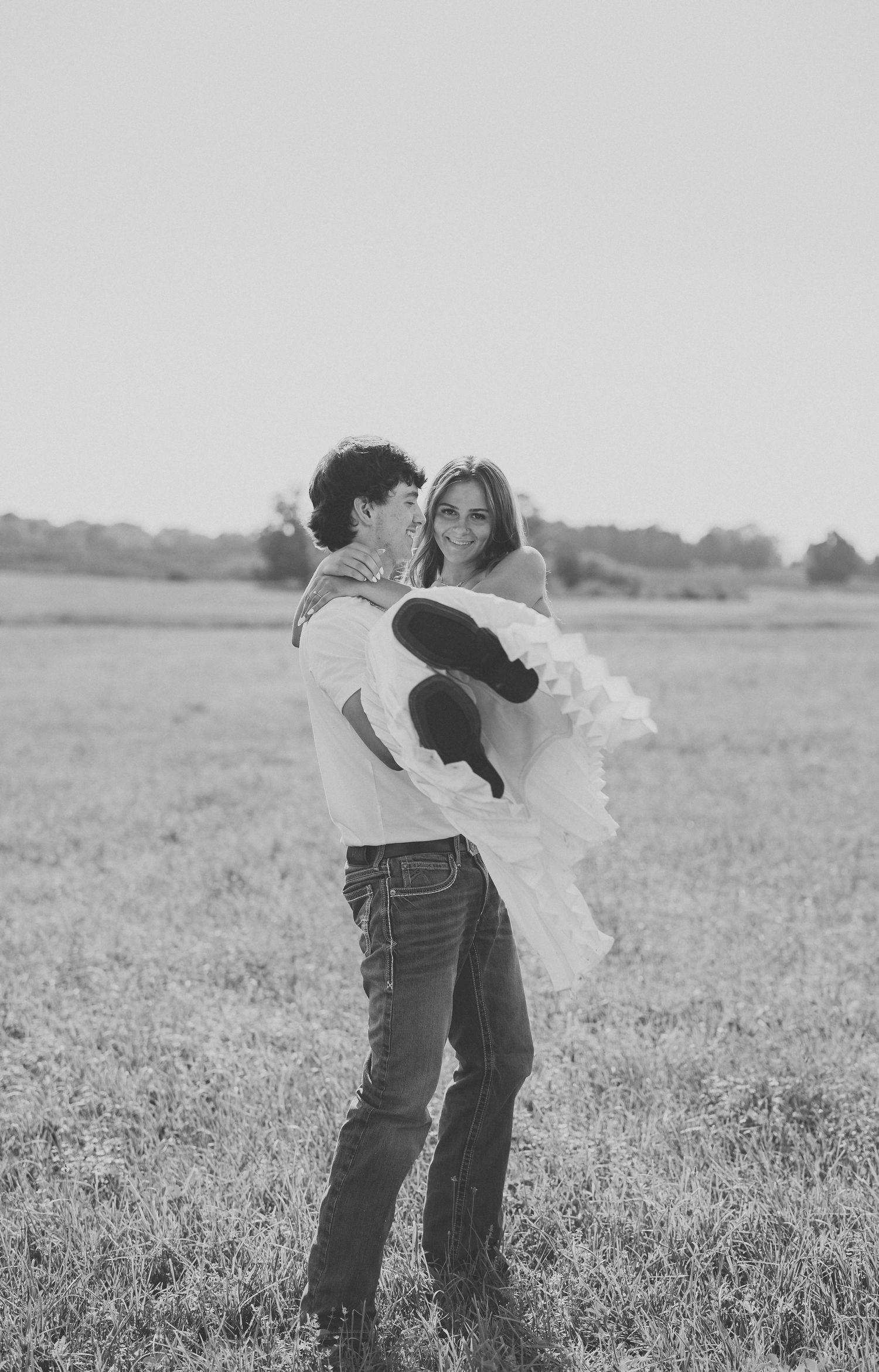 Black and white photo of a young couple in a field, man lifting woman in a white dress, smiling romantically.