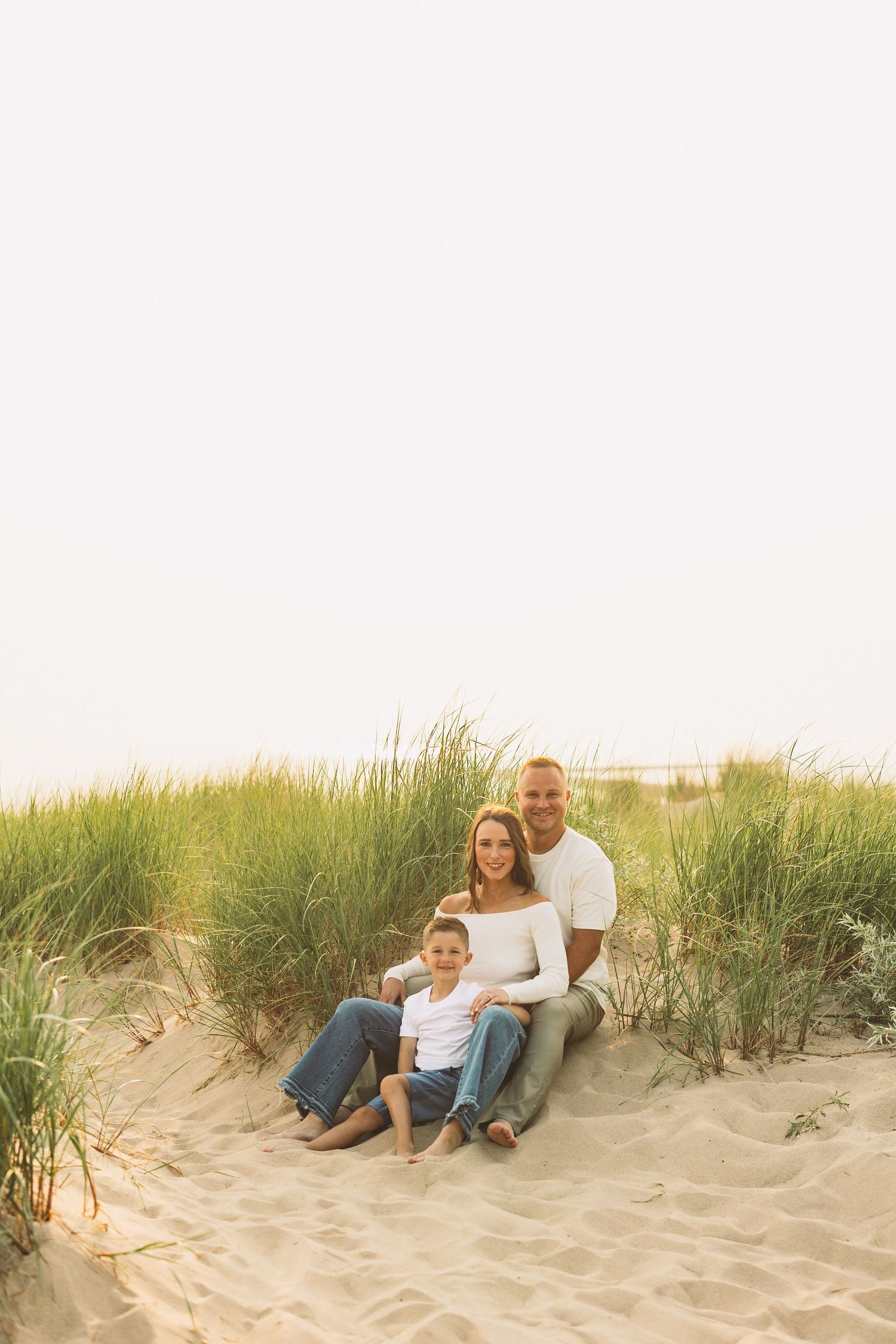 Family of three sitting on sandy beach dunes with green grass, smiling together in casual white and blue outfits.