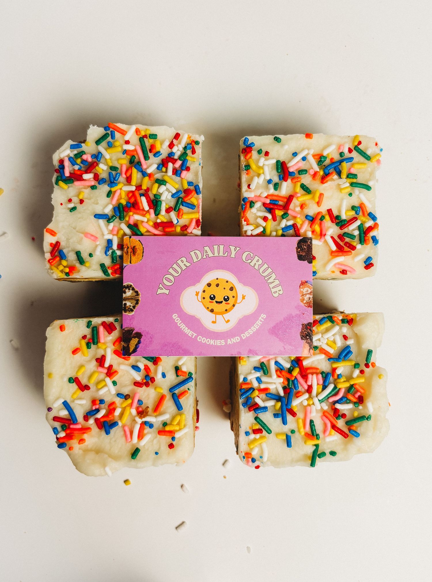 Four frosted birthday cake bars topped with rainbow sprinkles arranged around a Your Daily Crumb bakery box.