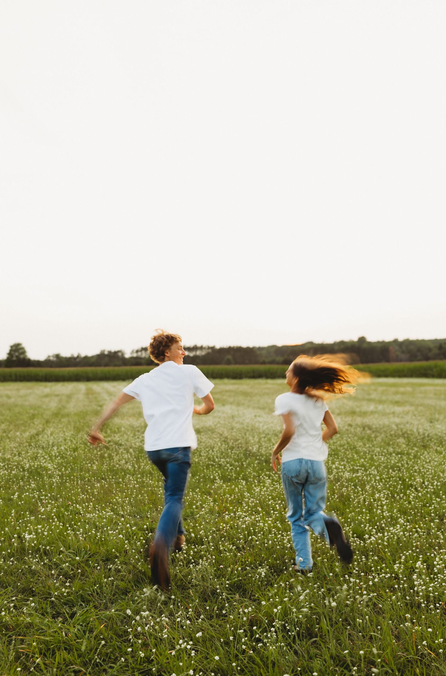 Two children in white shirts and jeans running through a lush green flower field at dusk.