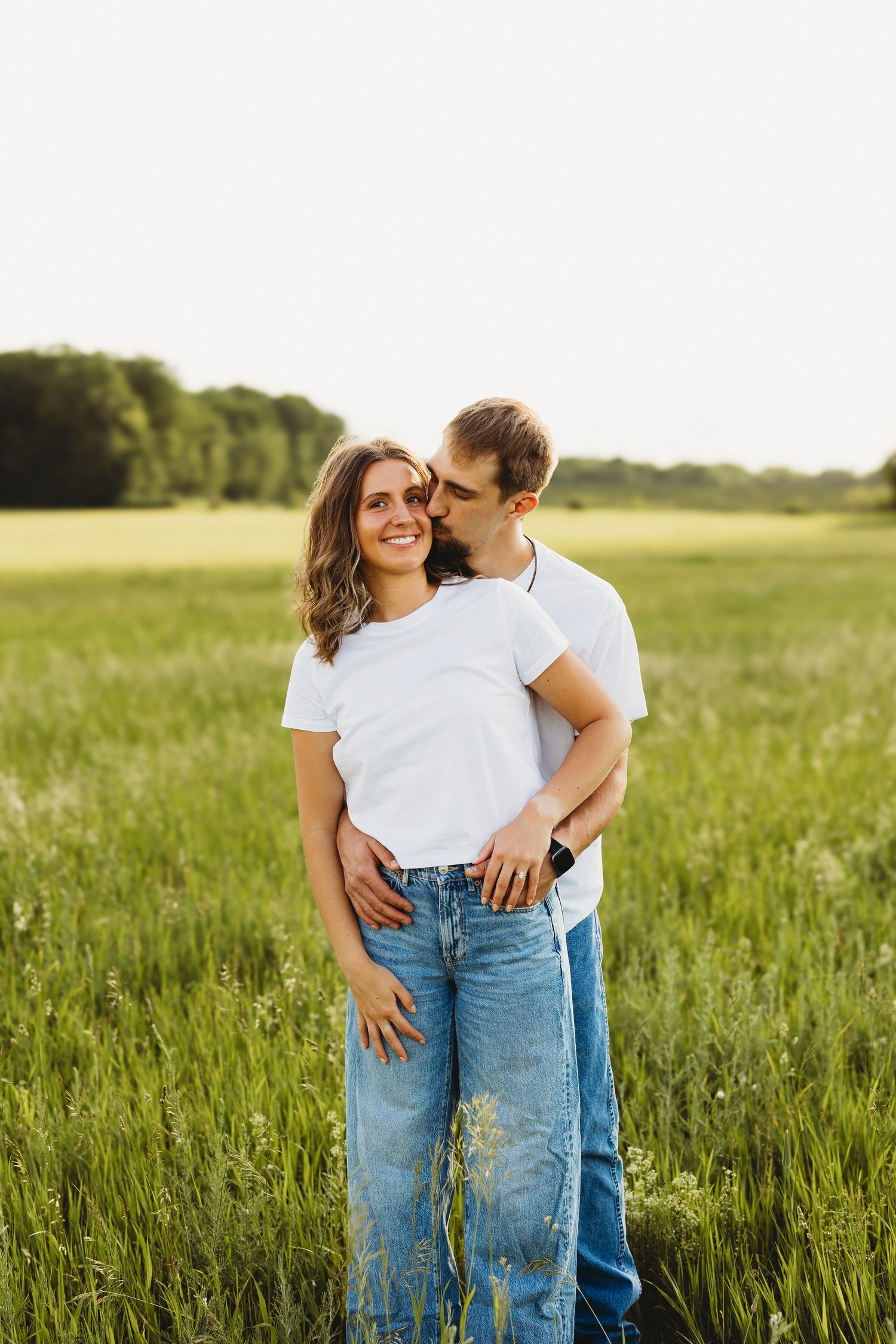 Couple in white t-shirts and jeans embracing in a green field, man kissing woman on cheek at sunset.