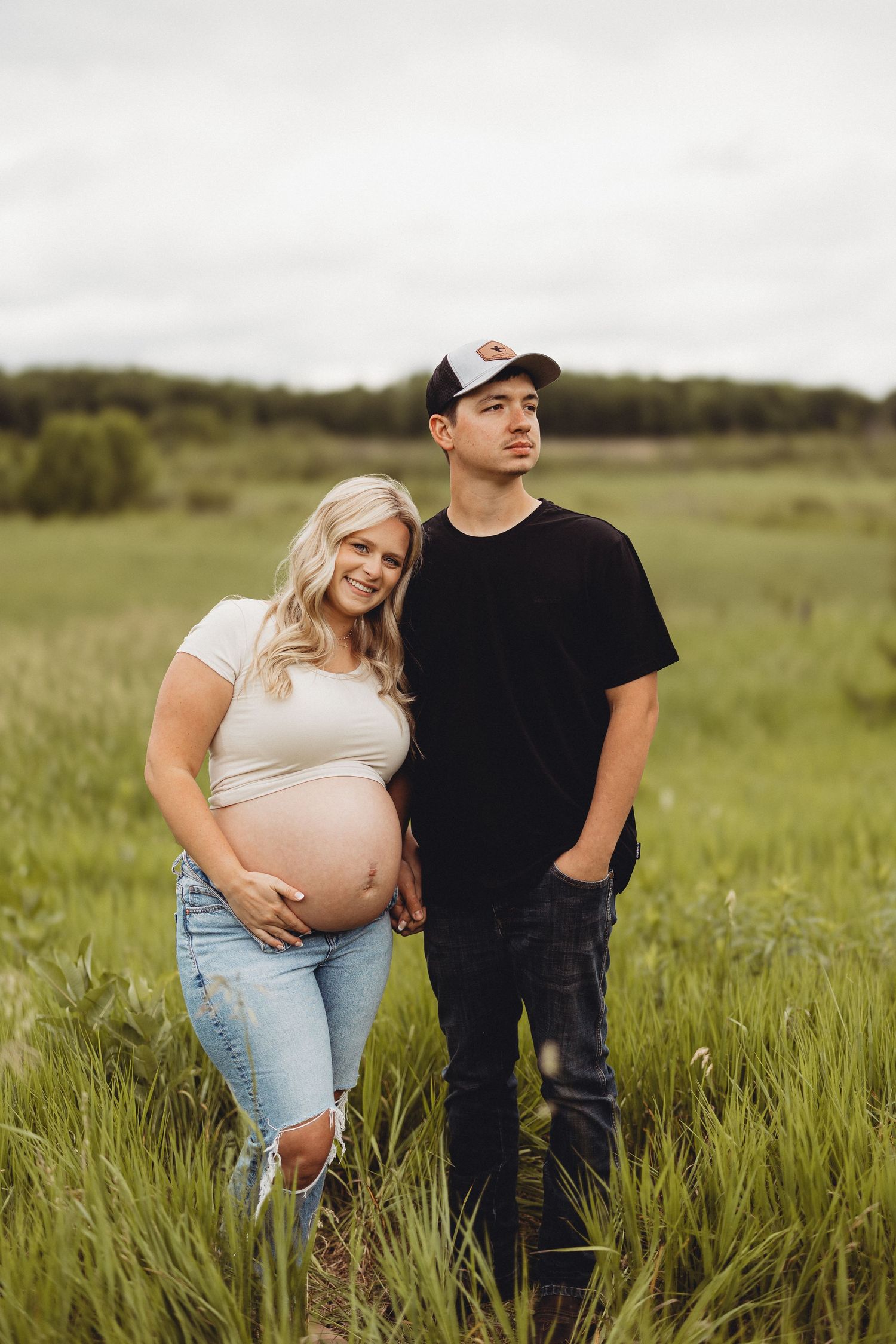 Pregnant couple standing in green field, woman in white top and jeans.