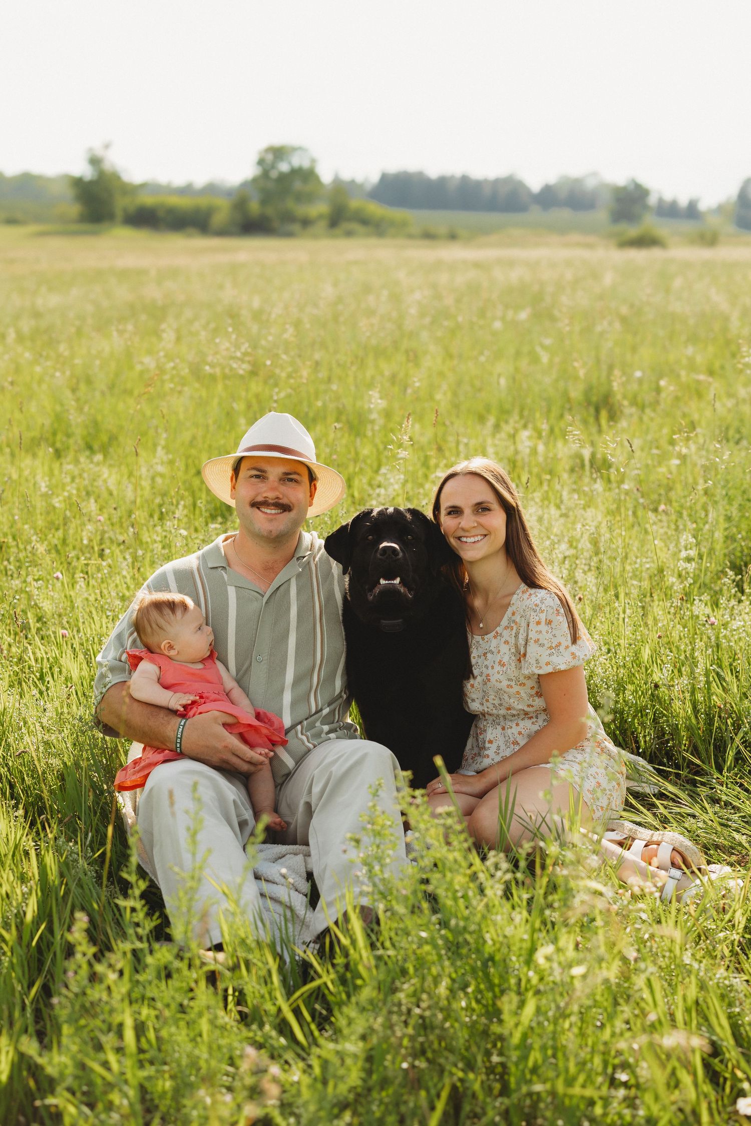 A smiling family with a baby and black dog sitting in a lush green field on a sunny day.