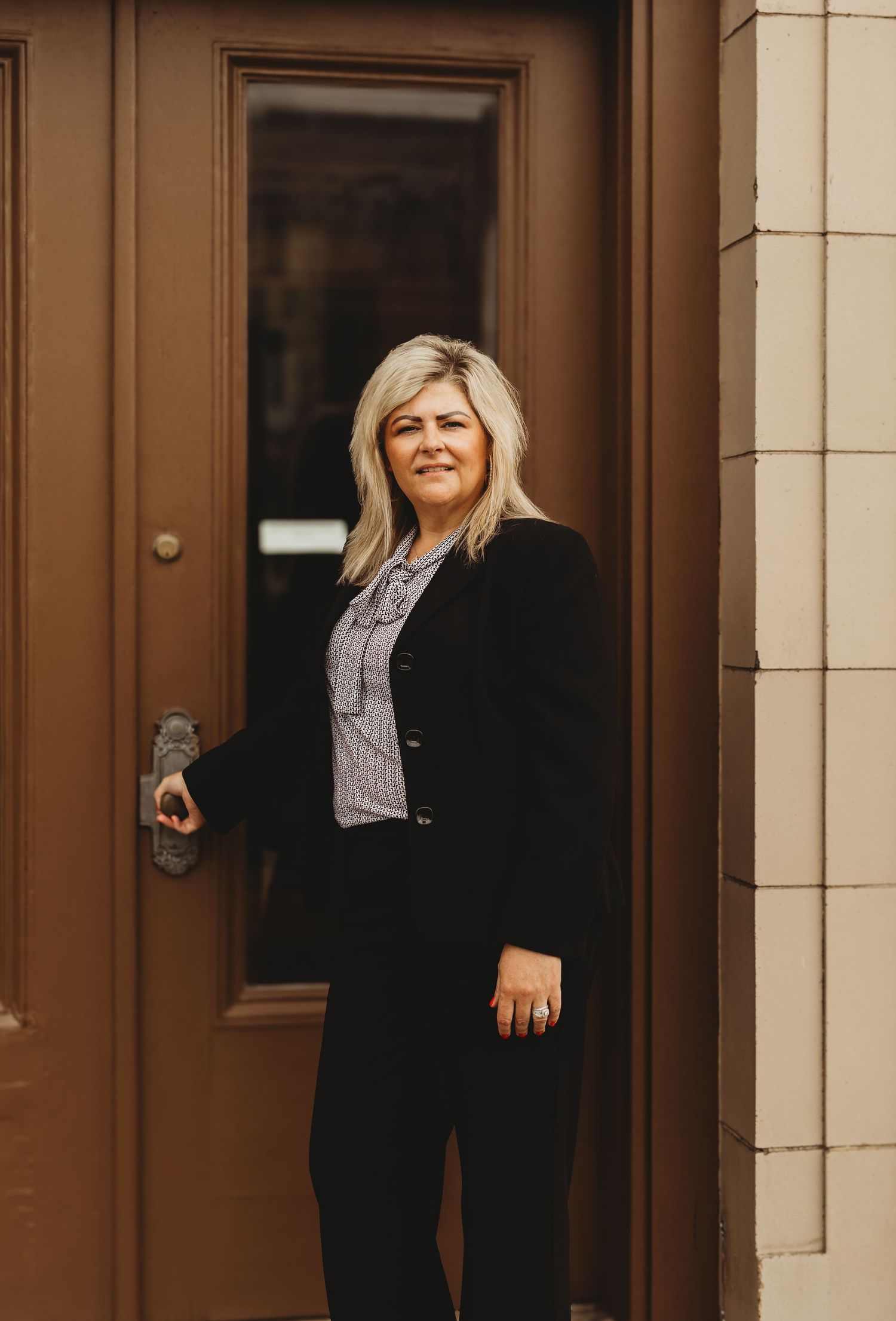 Professional blonde woman in black blazer standing by brown door in business attire.