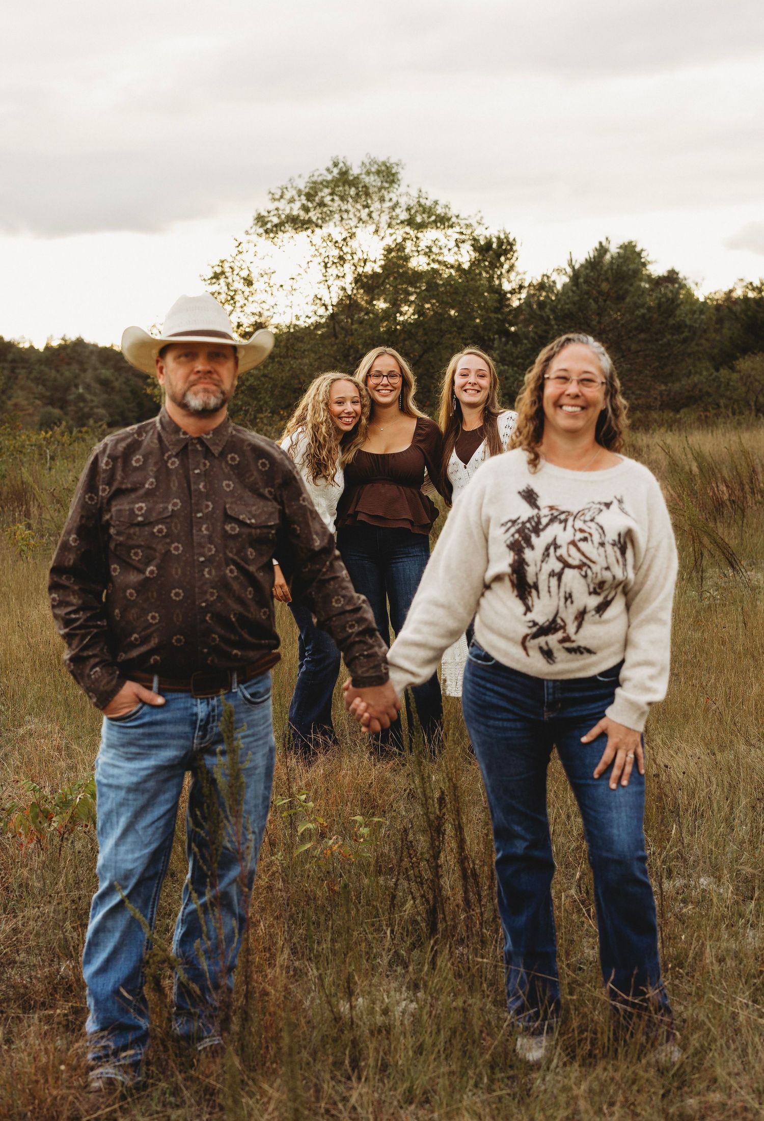 Family of five holding hands and walking through a tall grassy field at sunset, with a man in a cowboy hat and jeans leading the group.