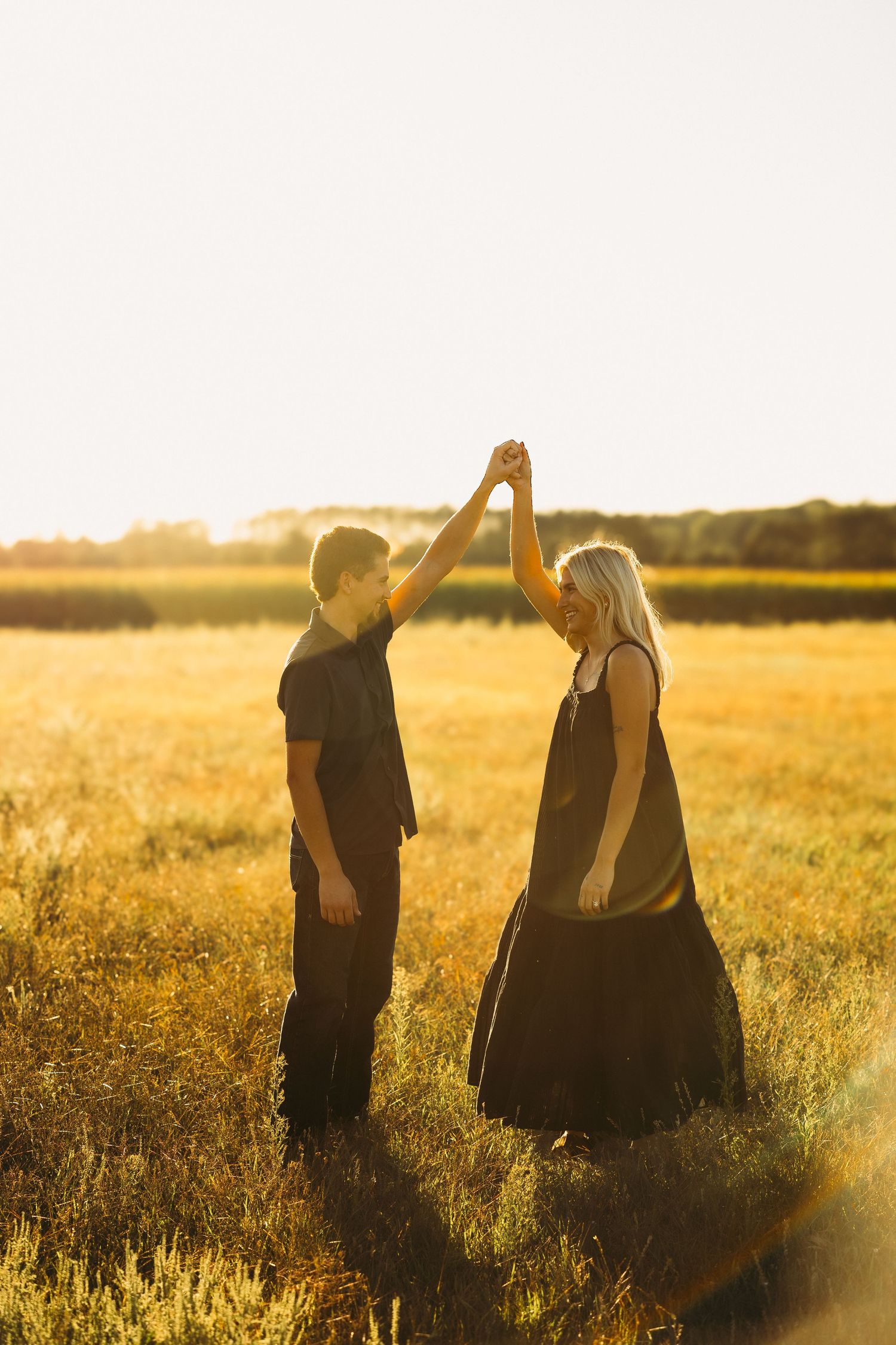 Couple holding raised hands in a dance pose in a golden grass field at sunset, both dressed in dark clothing.