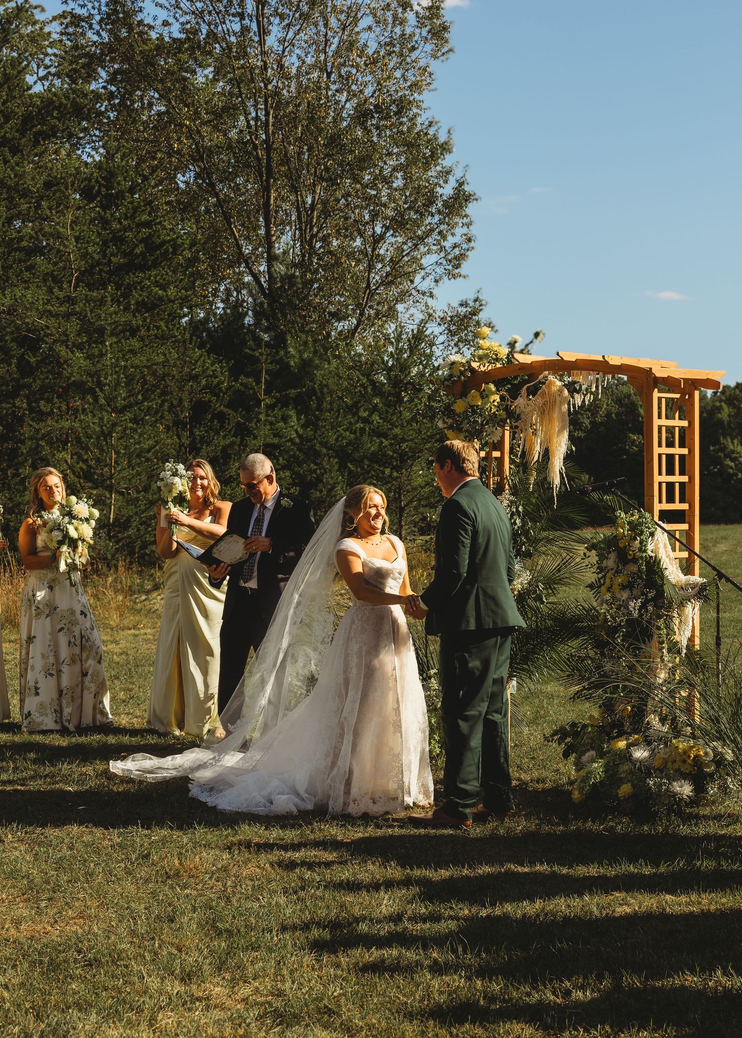 Bride in a flowing white gown and long veil holds hands with the groom during an outdoor wedding ceremony beneath a wooden arch.