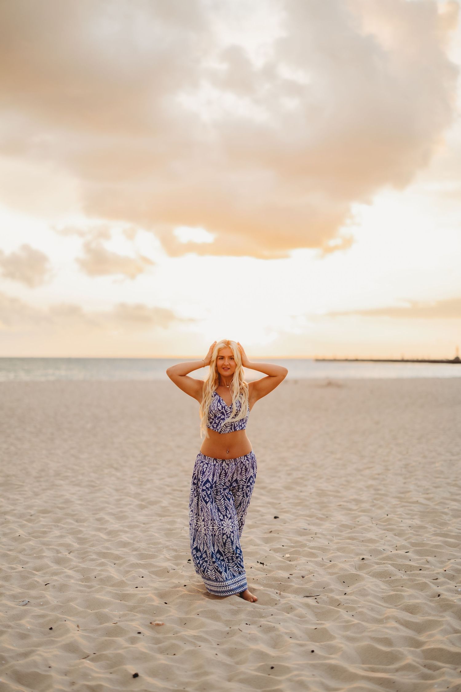 Blonde woman in a blue and white patterned two-piece outfit poses with hands in her hair on a sandy beach at golden sunset.