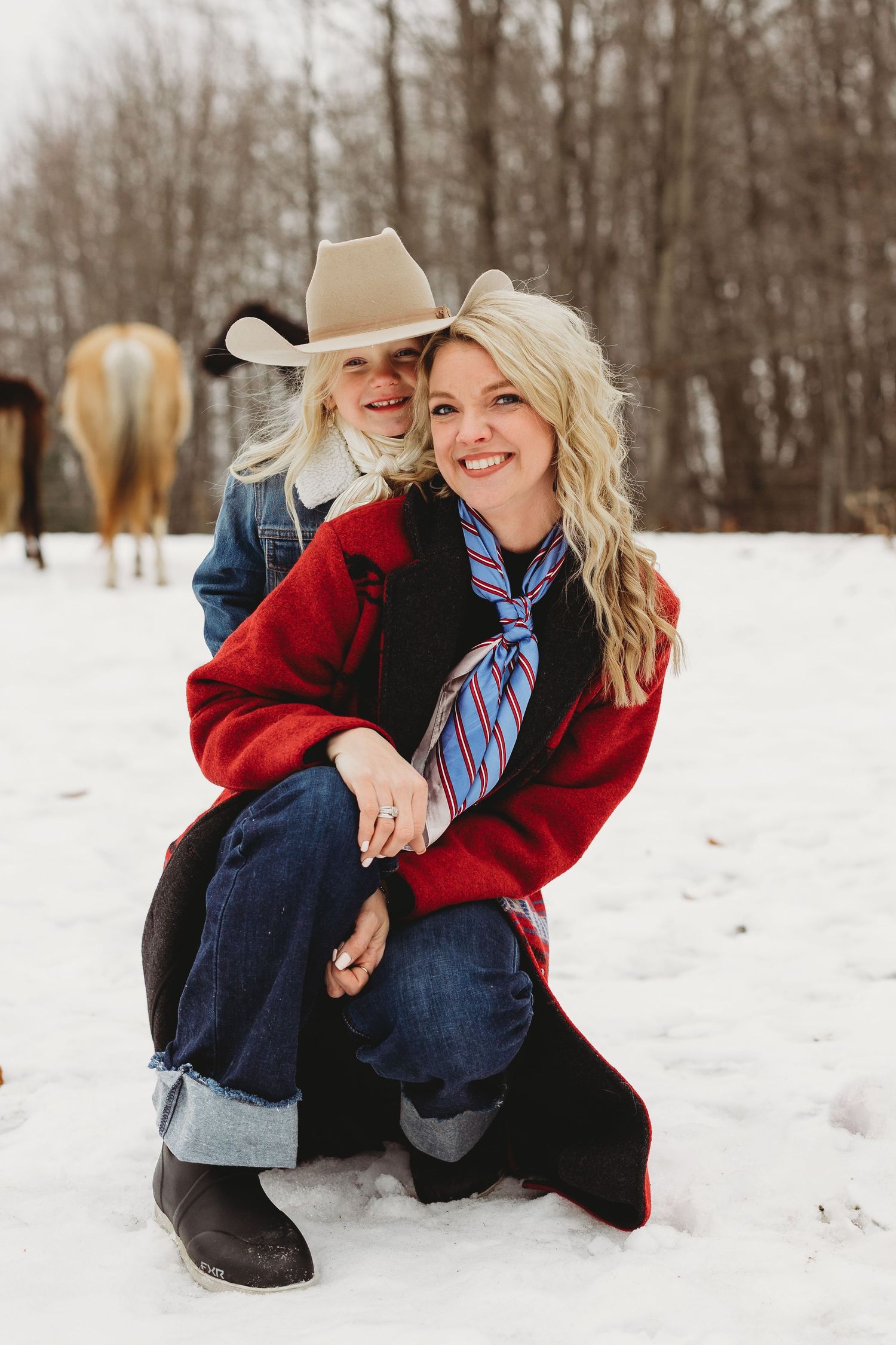 Woman in red coat crouching in snow with child in cowboy hat, horses visible in winter background.