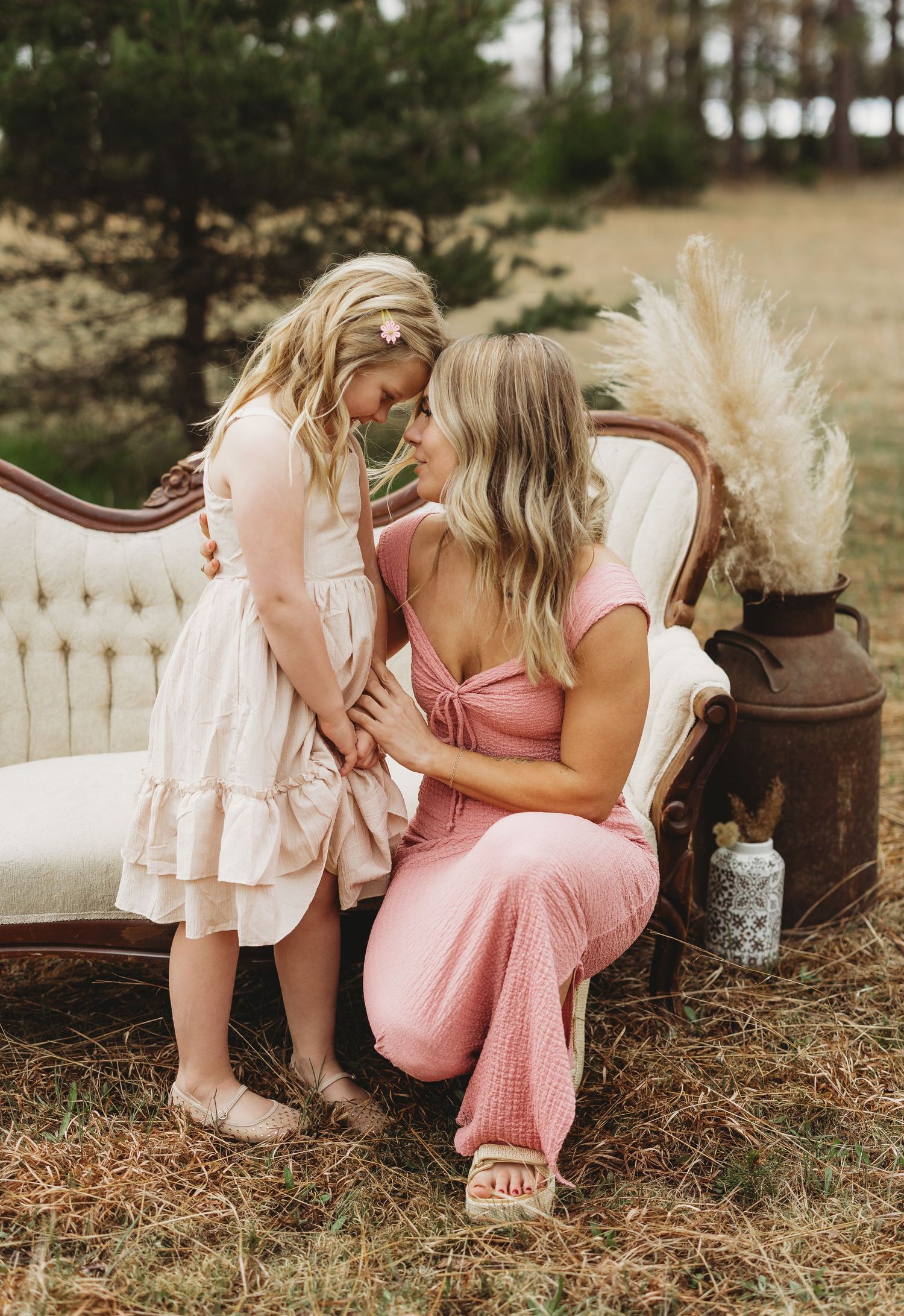 A mother in a pink jumpsuit kneels to share a tender moment with her young blonde daughter in a white dress outdoors.
