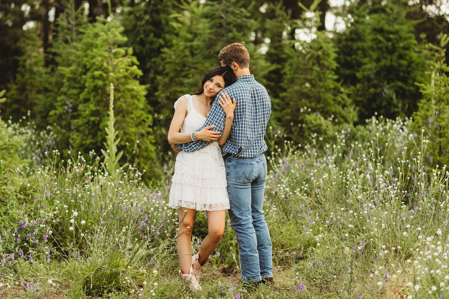 A couple embraces romantically in a lush wildflower meadow surrounded by tall green pine trees.