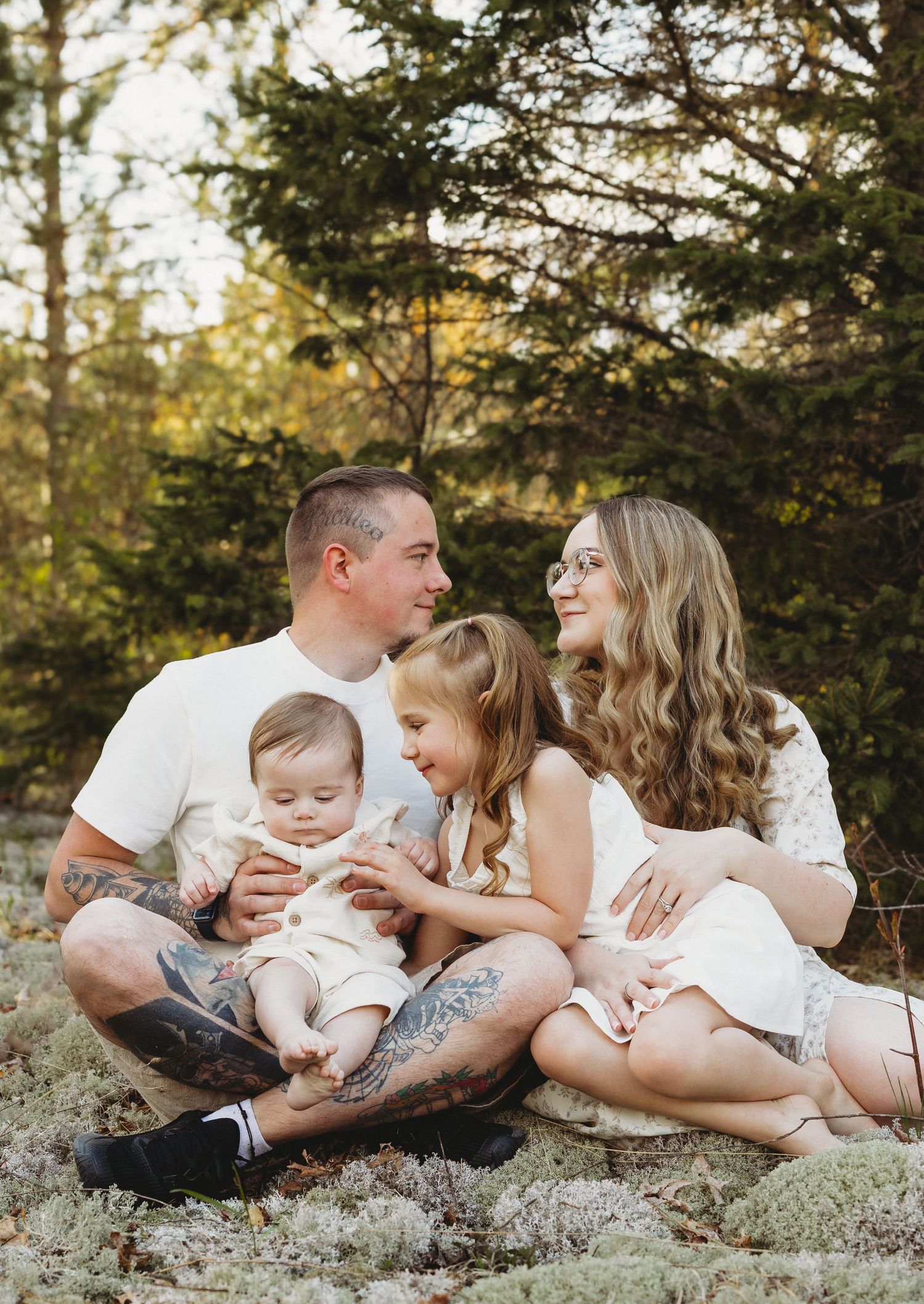 Family of four sitting outdoors in forest, parents with tattooed arms holding toddler and baby in white outfits.