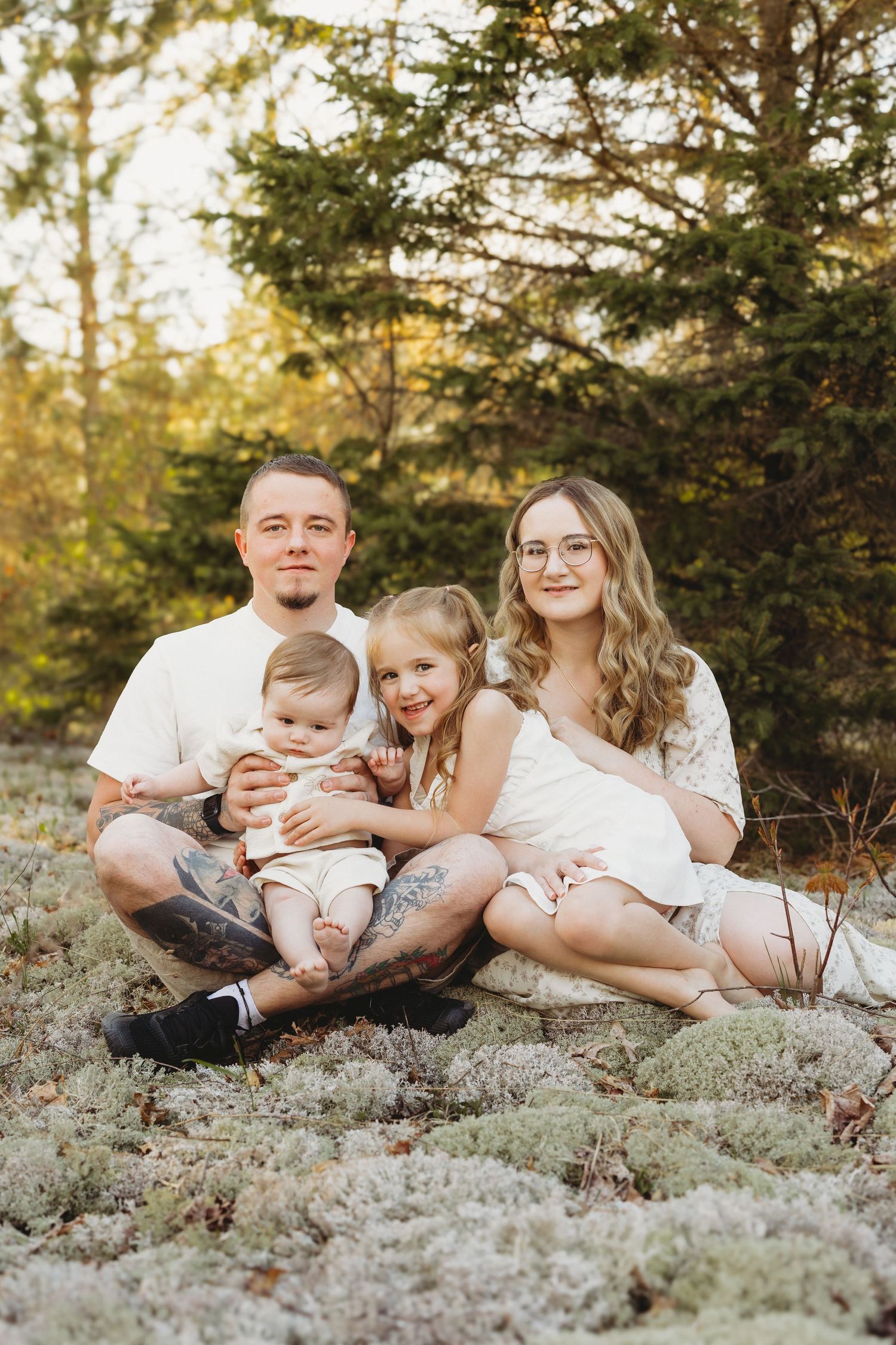 Happy family of four sitting outdoors in nature, parents with young daughter and baby smiling for a portrait photo.