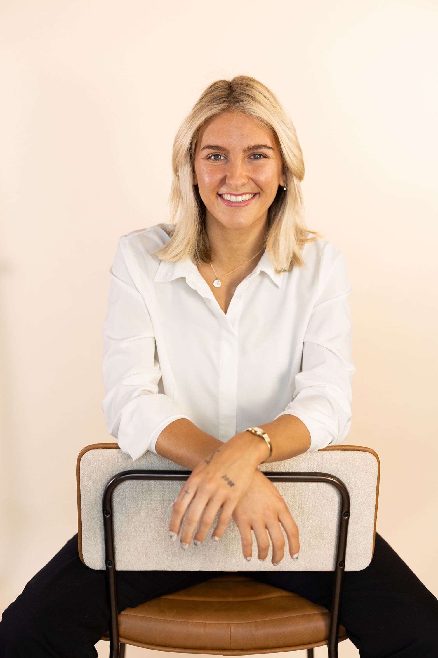 Smiling blonde woman in white shirt seated on chair with hands folded, professional headshot on beige background.