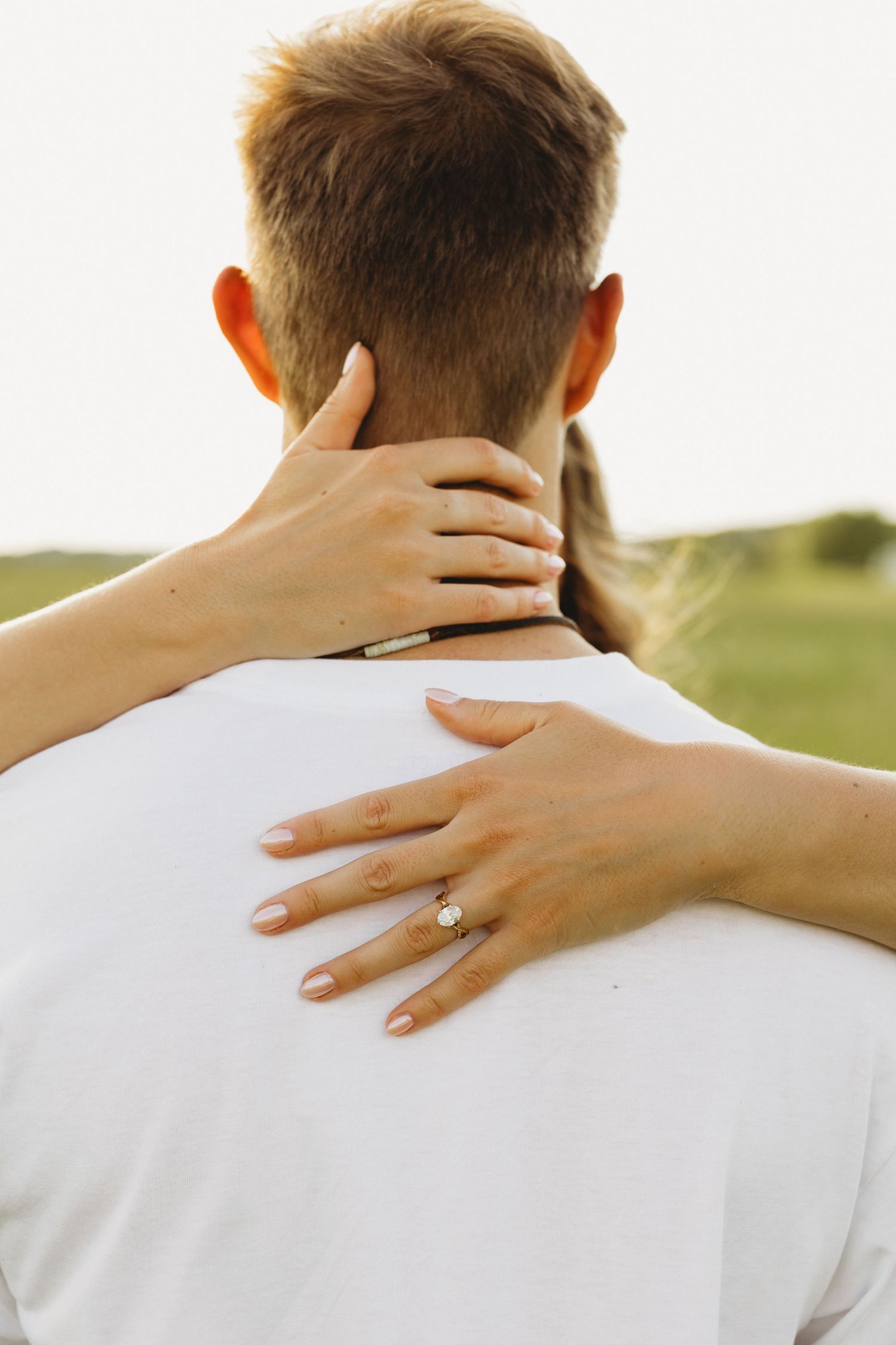 Close-up of woman's hands with engagement ring wrapped around man's neck outdoors.