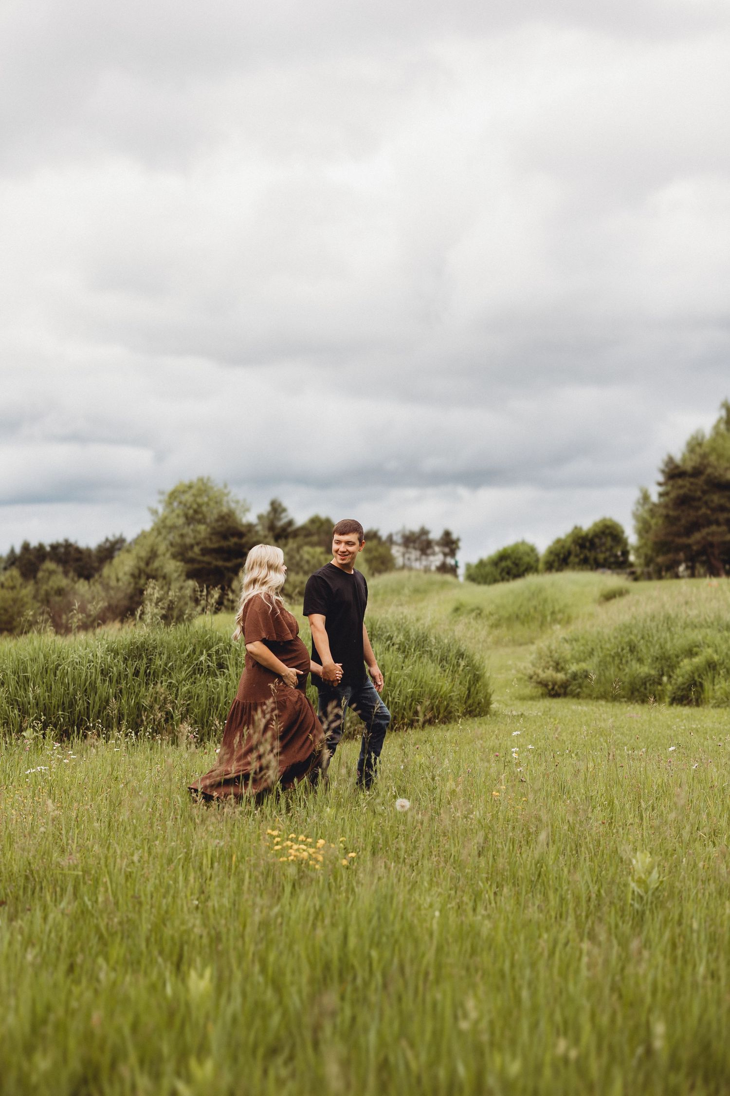 A couple holds hands walking through a lush green meadow, the woman in a brown dress and man in black.