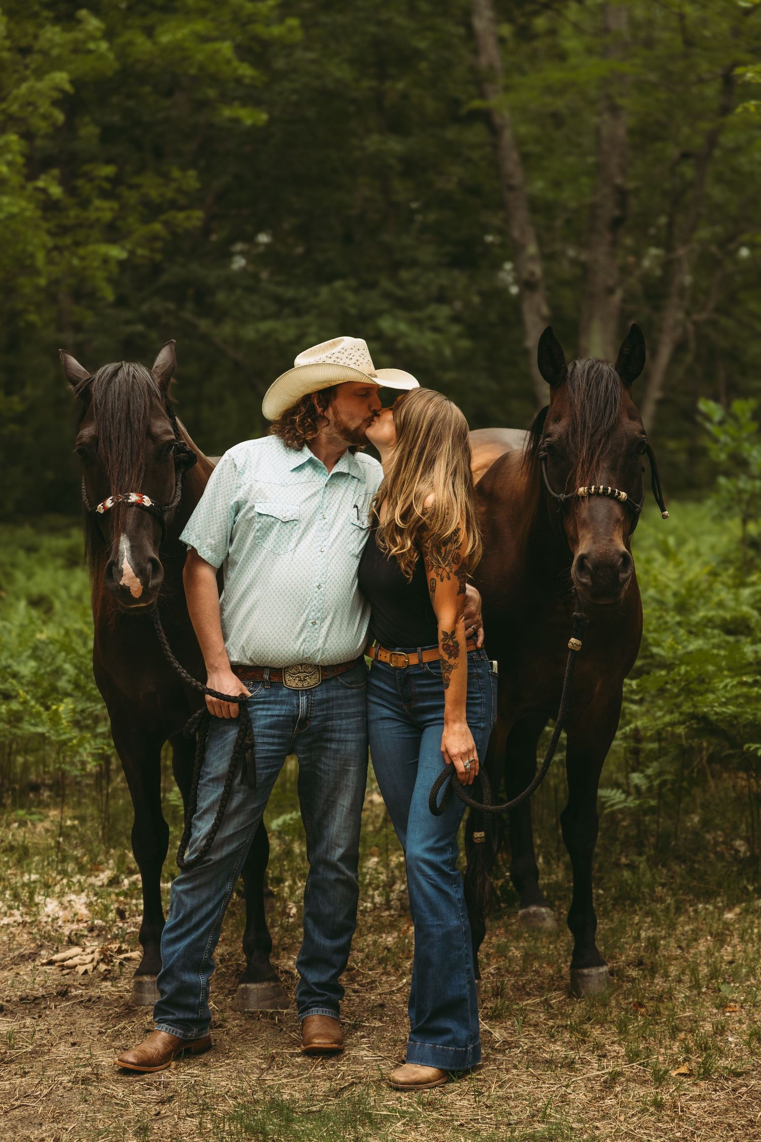 A couple sharing a kiss between two dark horses in a forest, man wearing cowboy hat and boots, woman in jeans.