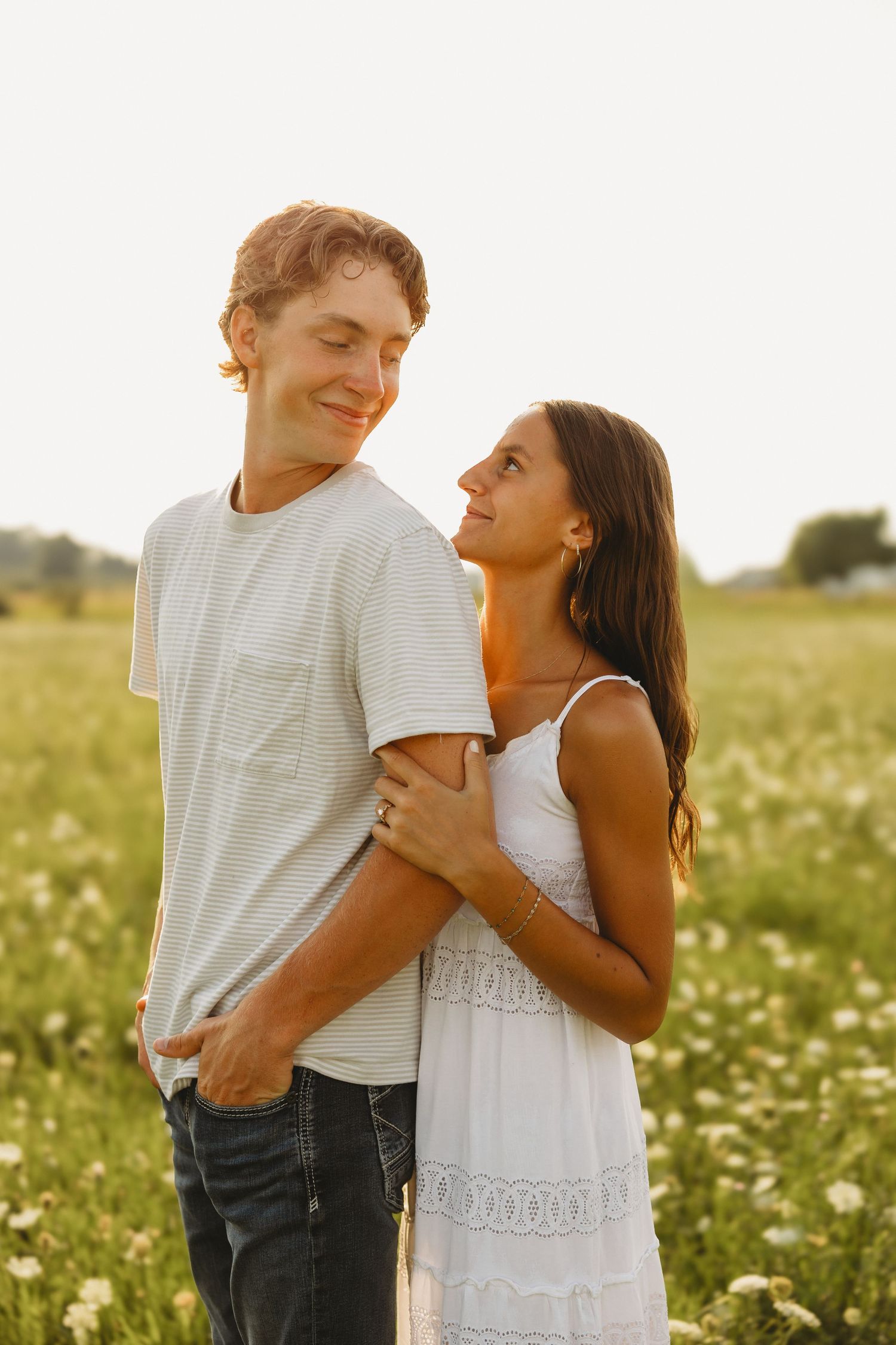 Couple embracing in a sunny flower field, woman in white dress gazing up at smiling man in striped tee at golden hour.