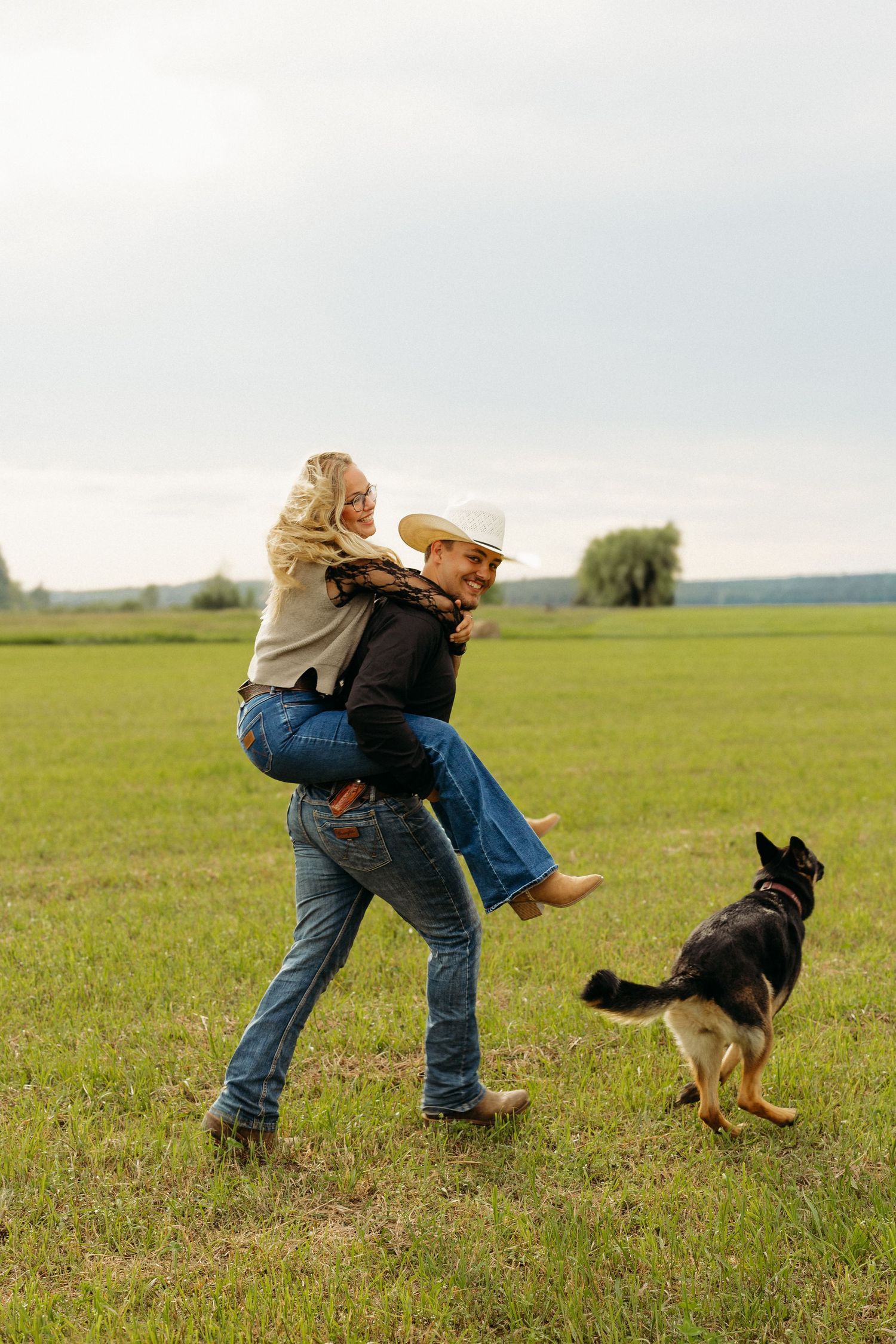 Man in cowboy hat giving woman piggyback ride in green field while German Shepherd dog plays nearby.