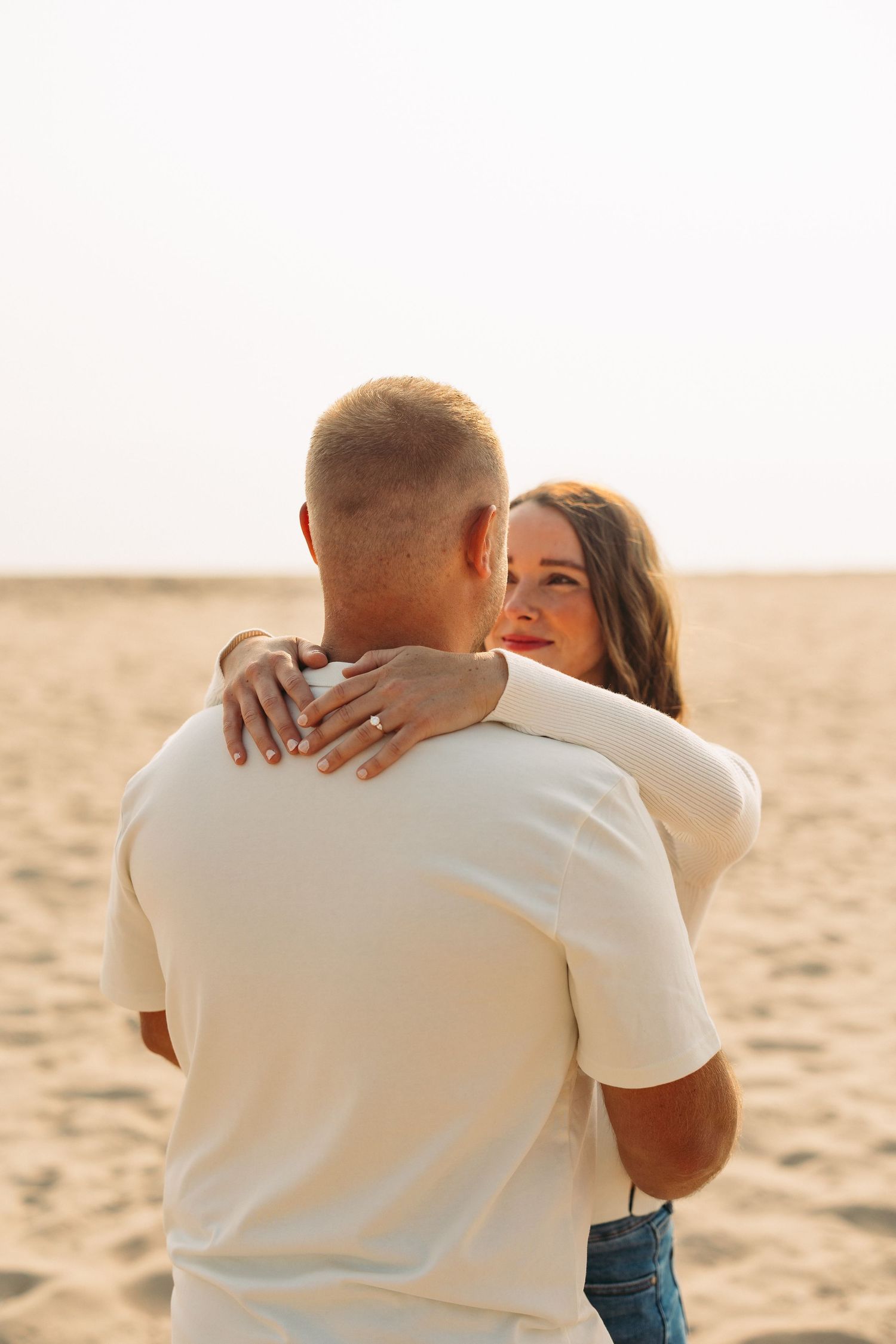 Couple embracing on a sandy beach, woman with red lips smiling, wearing a ring, man in white t-shirt.