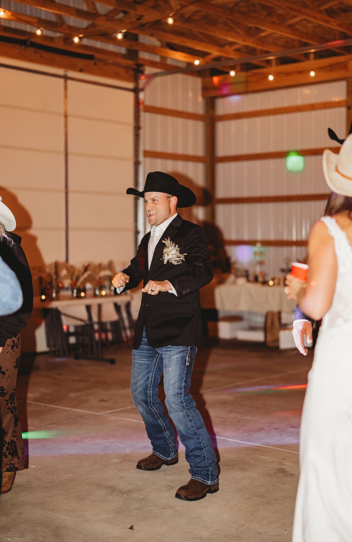 Groom in a black cowboy hat, blazer and jeans dances with a boutonniere at a rustic barn wedding reception while guests look on.
