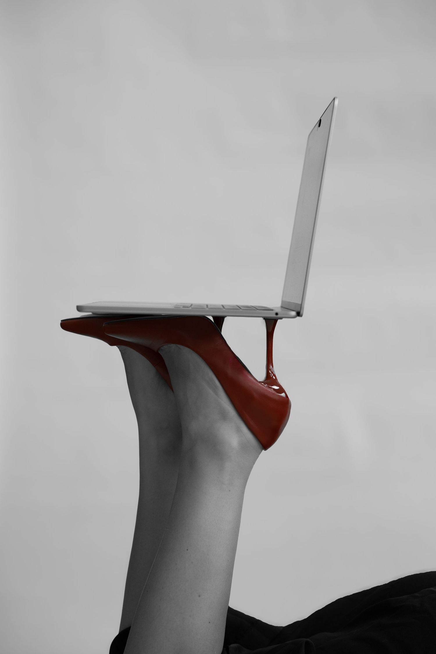 Woman's leg raised upward balancing an open silver laptop on a red high-heel shoe in a black and white photo with selective red color.