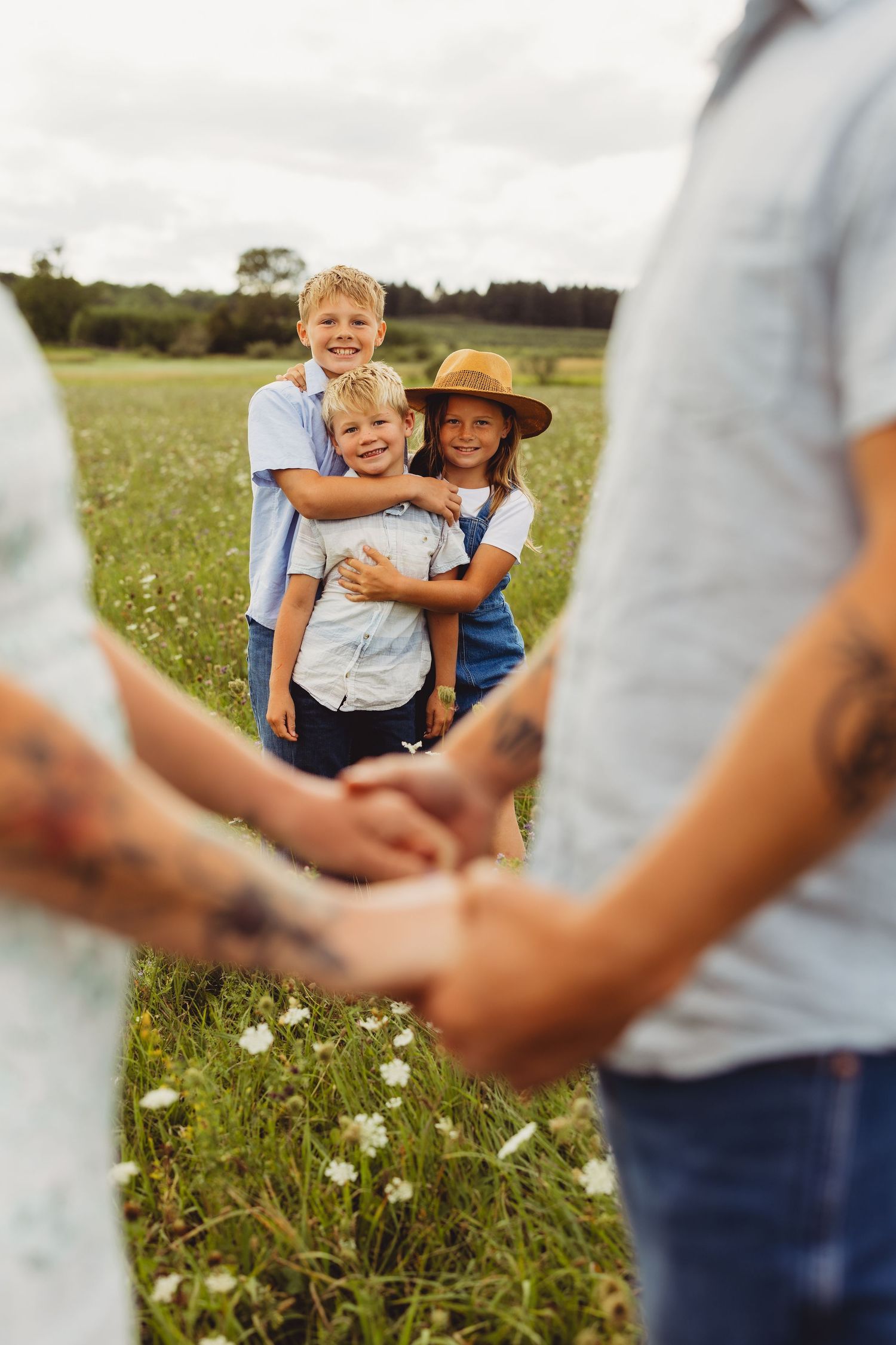 Three smiling children hug in a sunny wildflower meadow while two adults in the foreground hold hands in a family portrait.