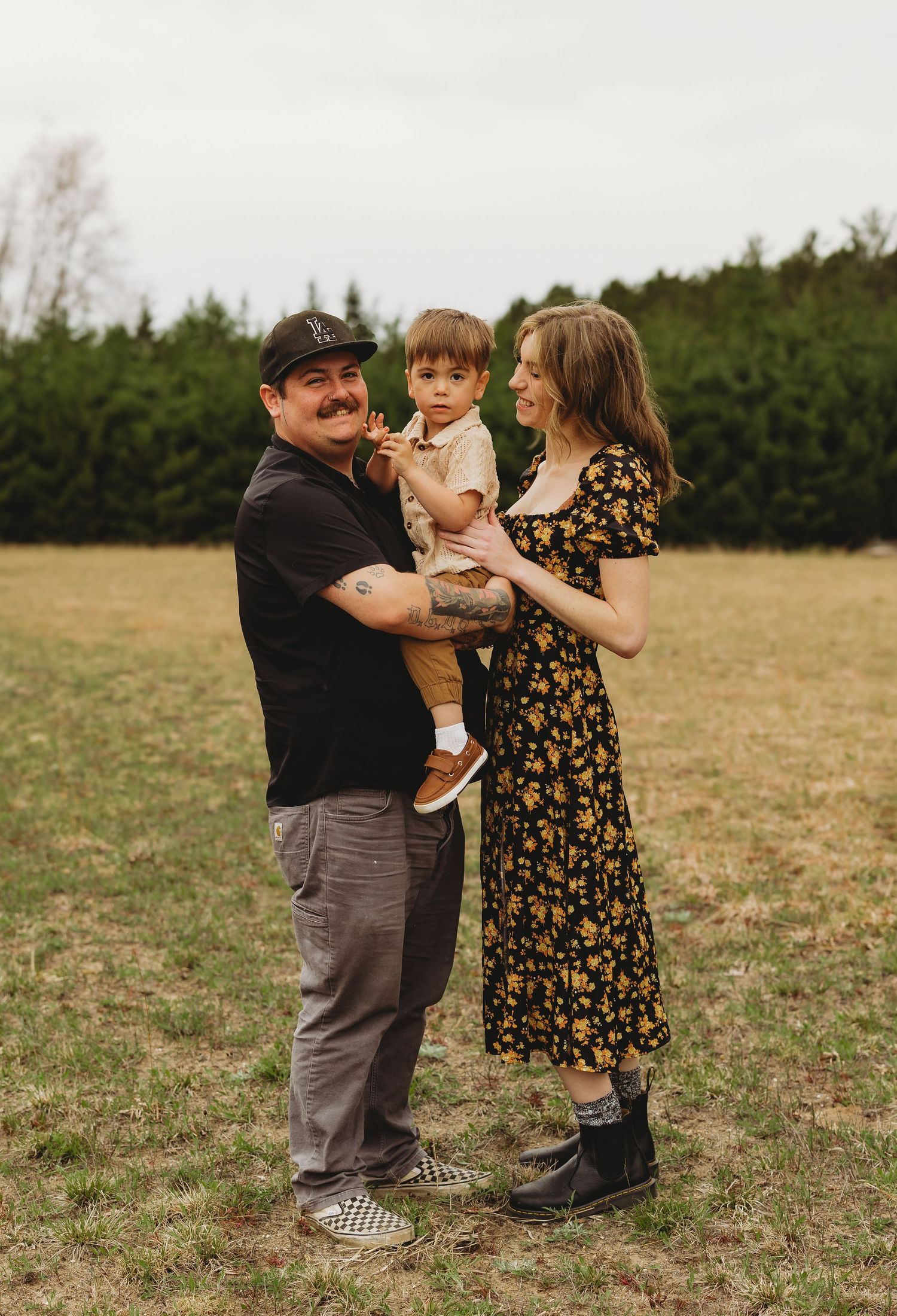 A couple stands in a grassy field holding their toddler, woman in floral dress, man in black shirt and cap.