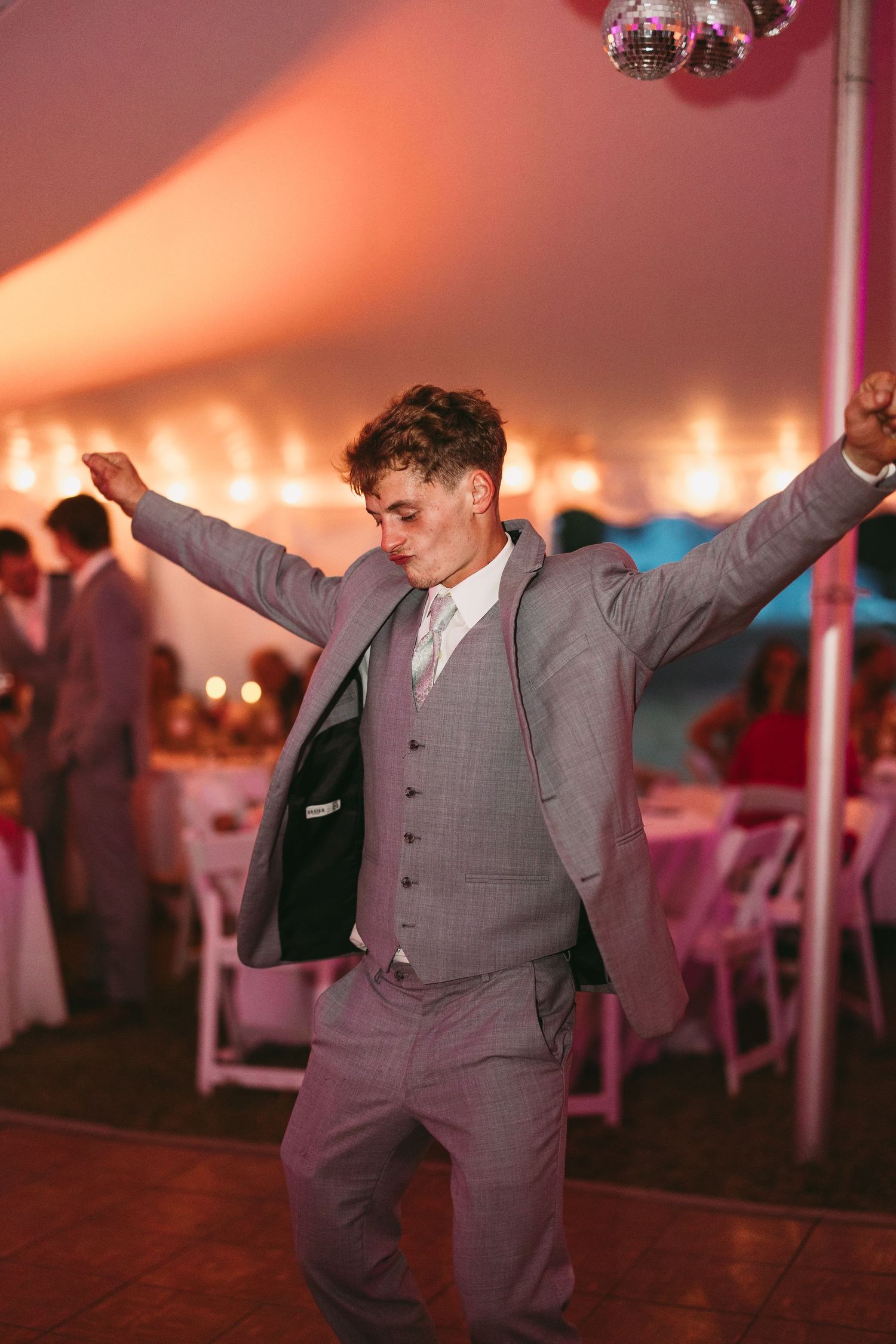 Wedding guest in grey suit dancing enthusiastically under warm lighting at reception.