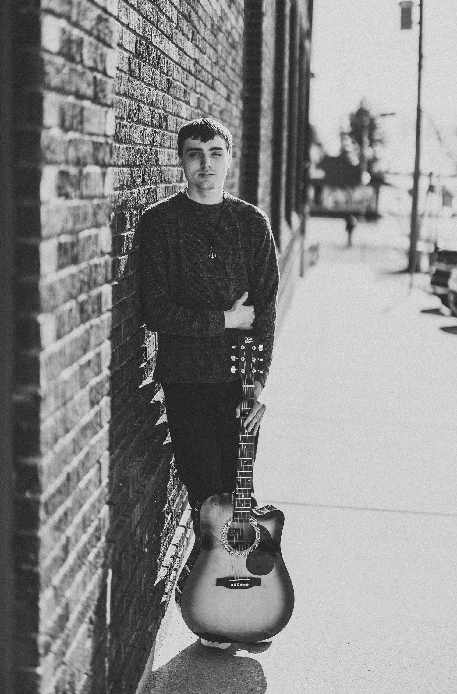 Musician in black clothing poses with acoustic guitar against brick wall in black and white photo series.
