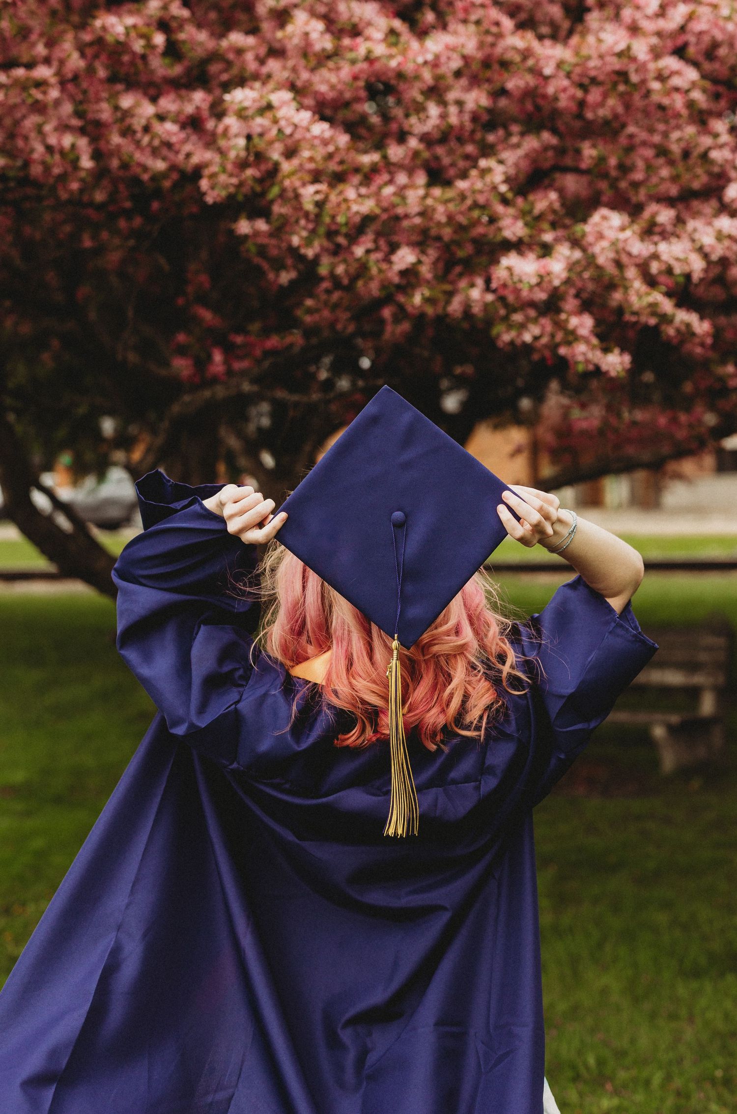 Graduate in blue cap and gown playfully poses under pink cherry blossoms.