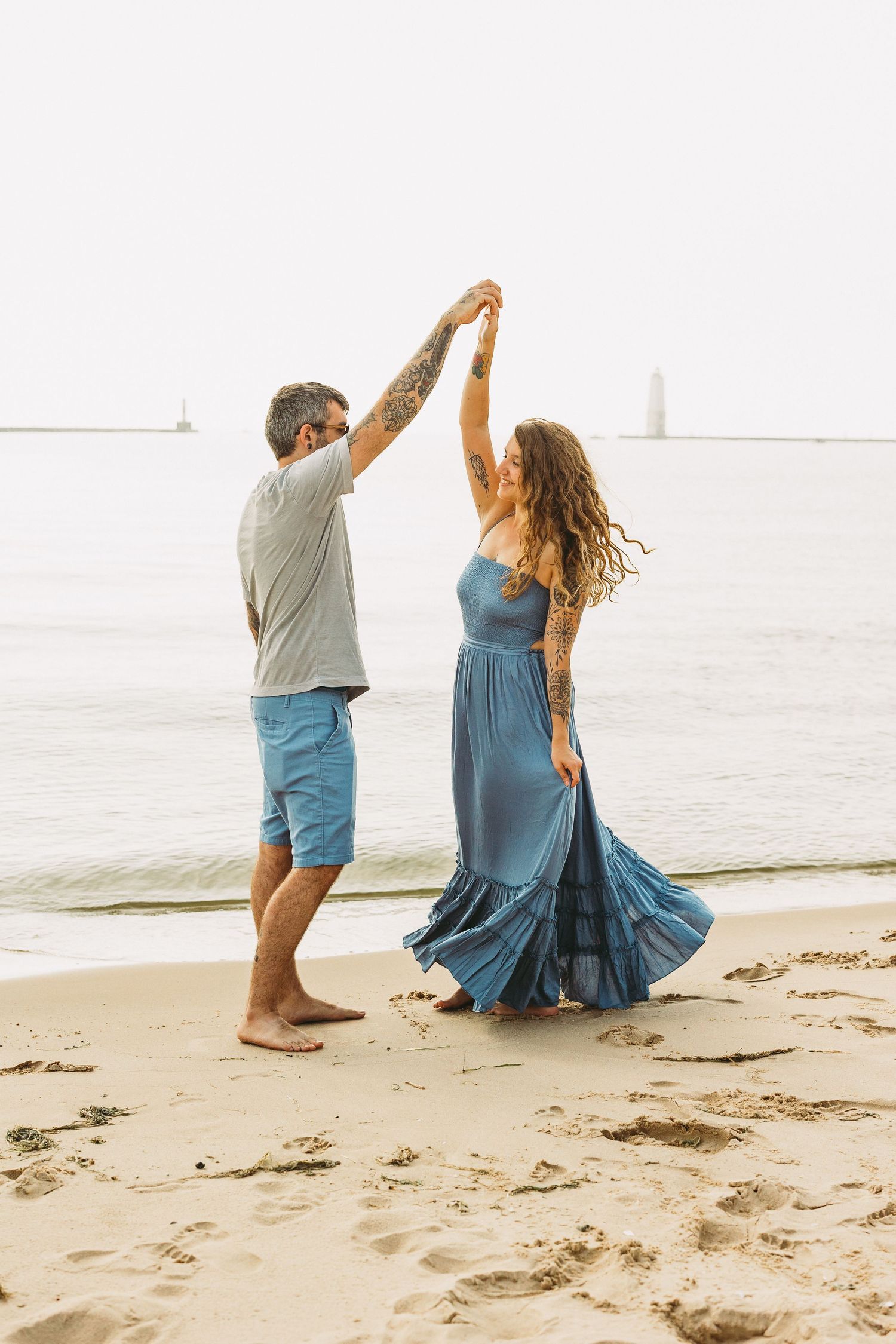 Couple in blue outfits dancing and twirling together on a sandy beach at sunset with ocean waves in background.