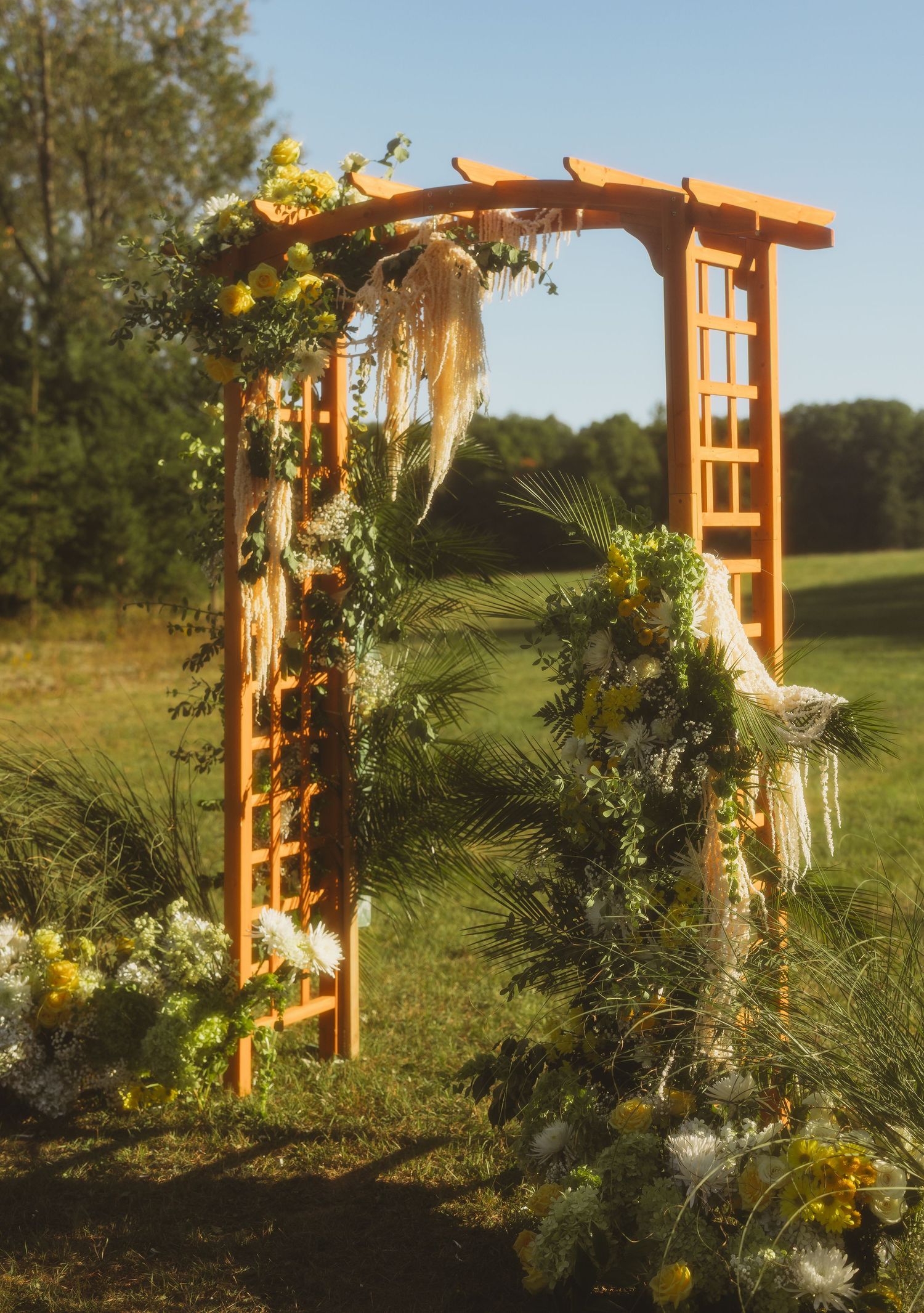 Wooden wedding arch decorated with yellow and white flowers, greenery and pampas grass, set in a sunny outdoor field with trees behind.