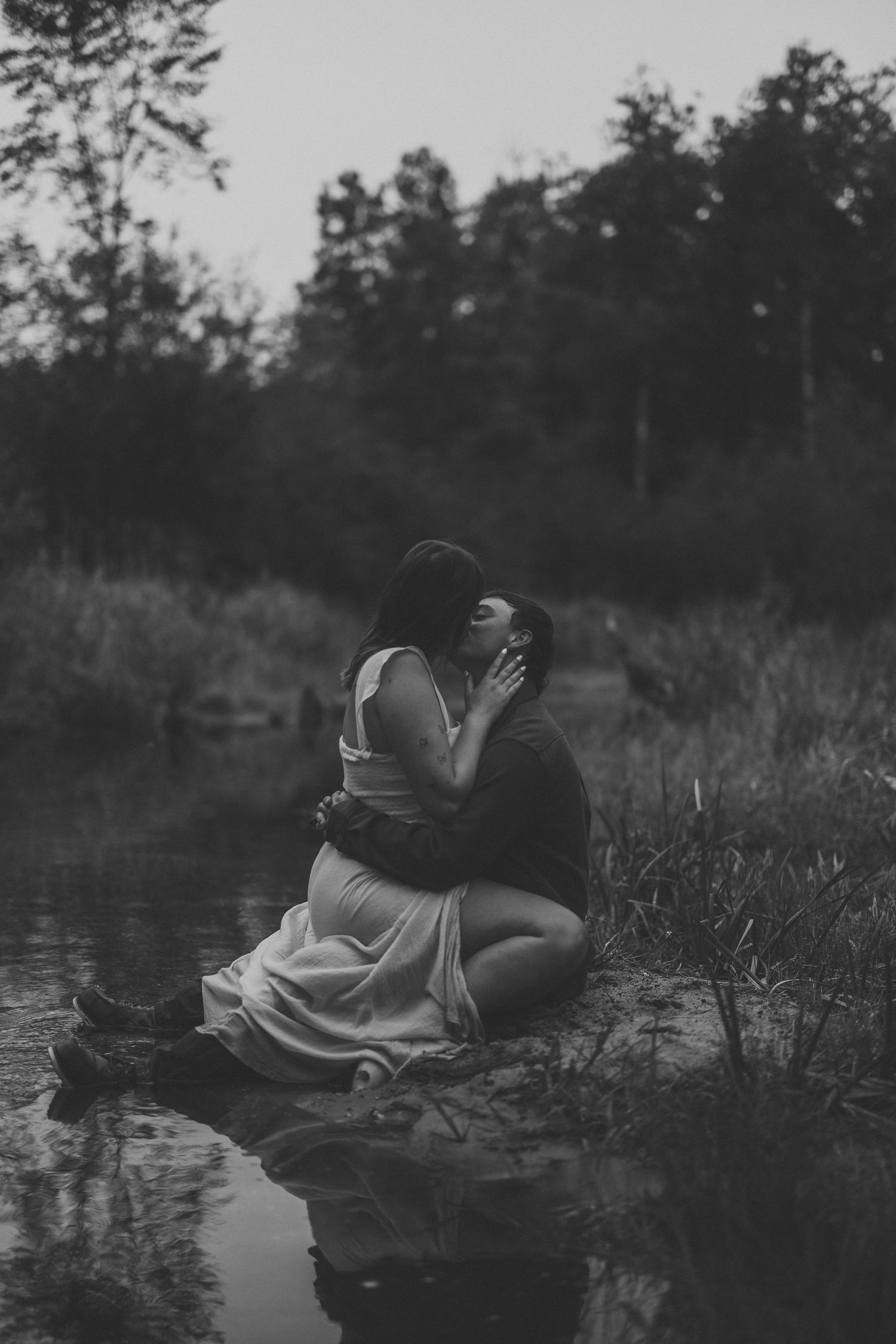 Black and white photo of a couple kissing while seated at the edge of a calm pond, surrounded by tall grass and trees.