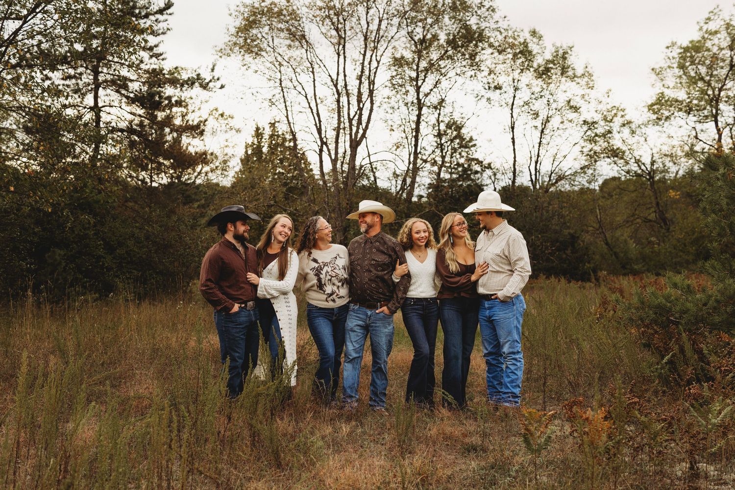 Group of eight family members in cowboy hats and western attire standing arm in arm in a grassy field with trees in the background.