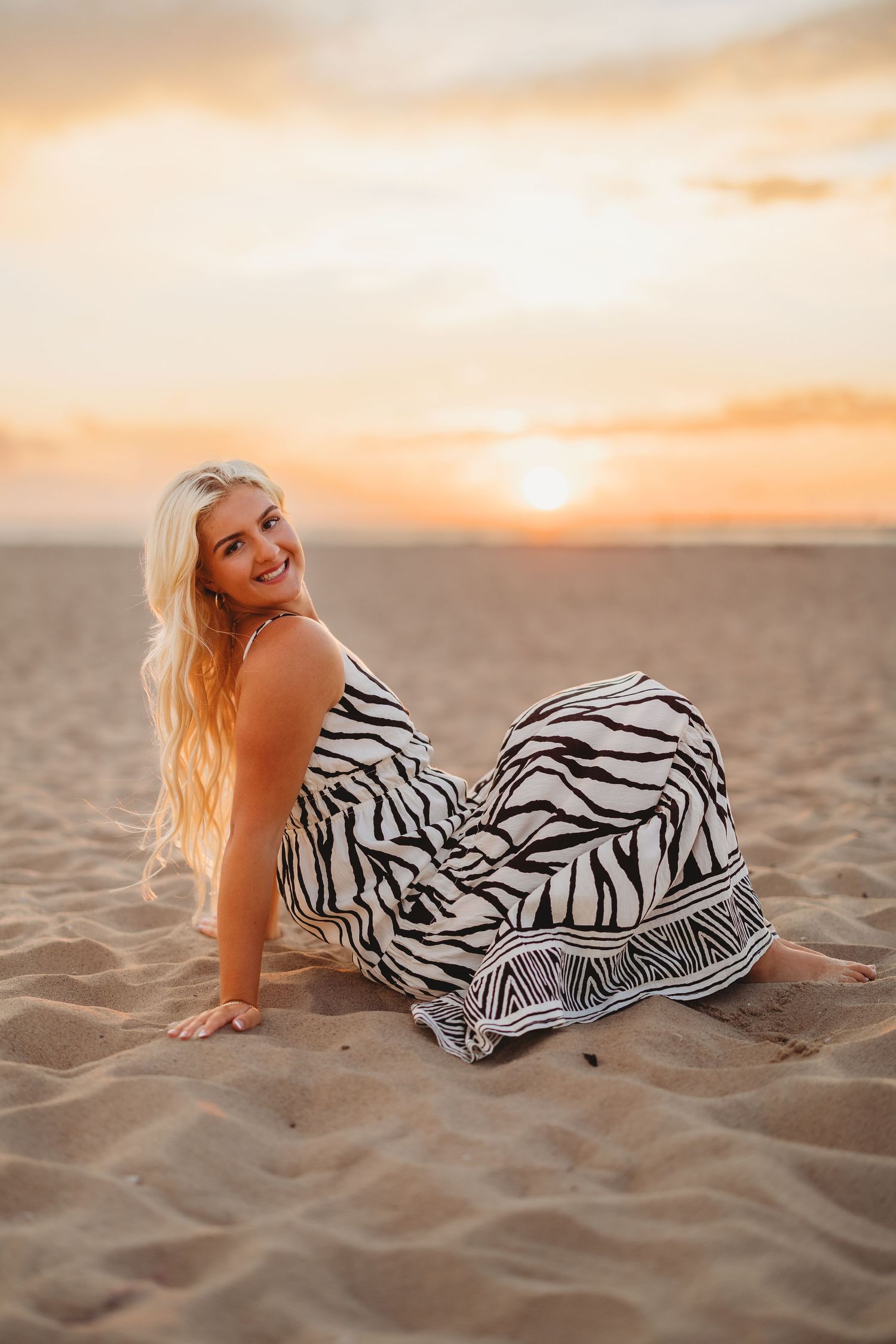 Smiling blonde woman in a black and white zebra print maxi dress sitting on the sand at the beach during a colorful sunset.