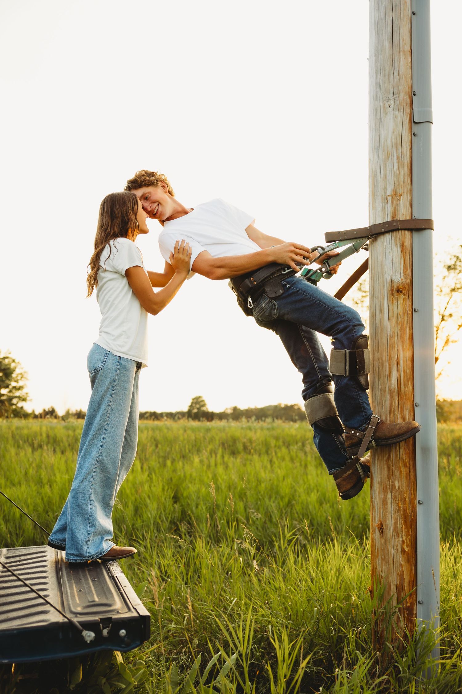 A young couple shares a romantic moment as a man climbs a wooden utility pole in a green field.