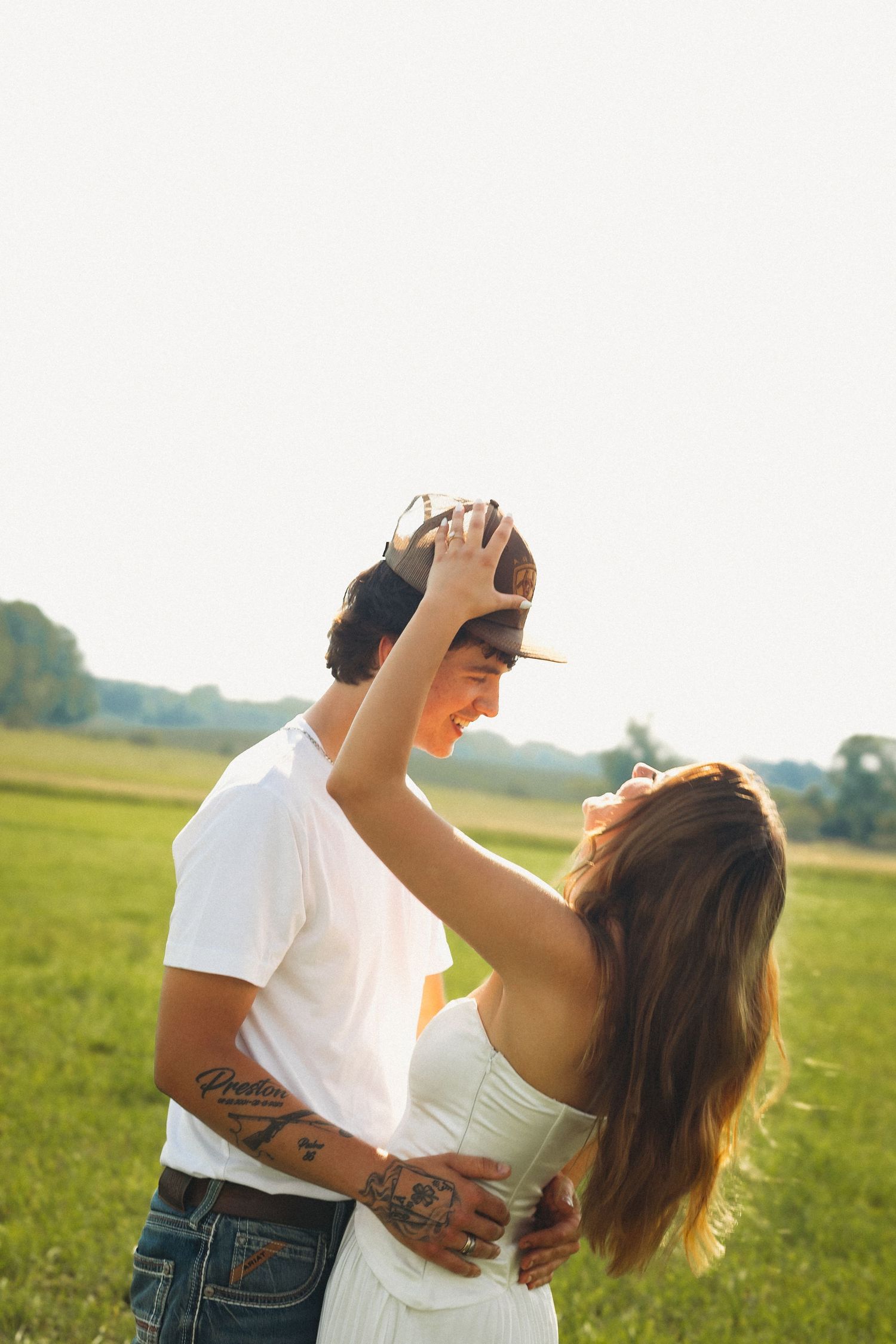 A couple embraces in a sunny green field, woman playfully adjusting mans cap, both smiling, tattoos visible.
