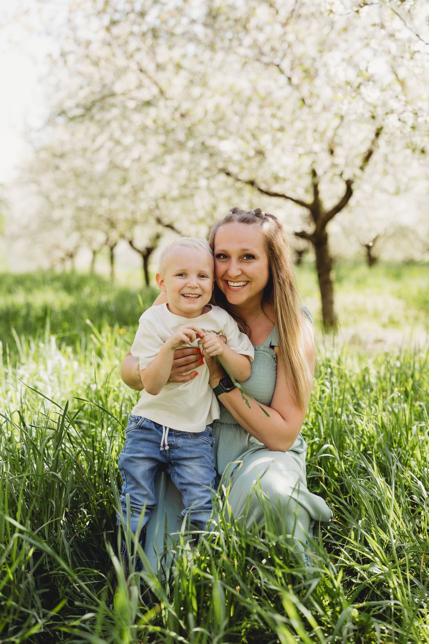 A smiling mother crouches in green grass holding her happy toddler son near blooming spring trees outdoors.