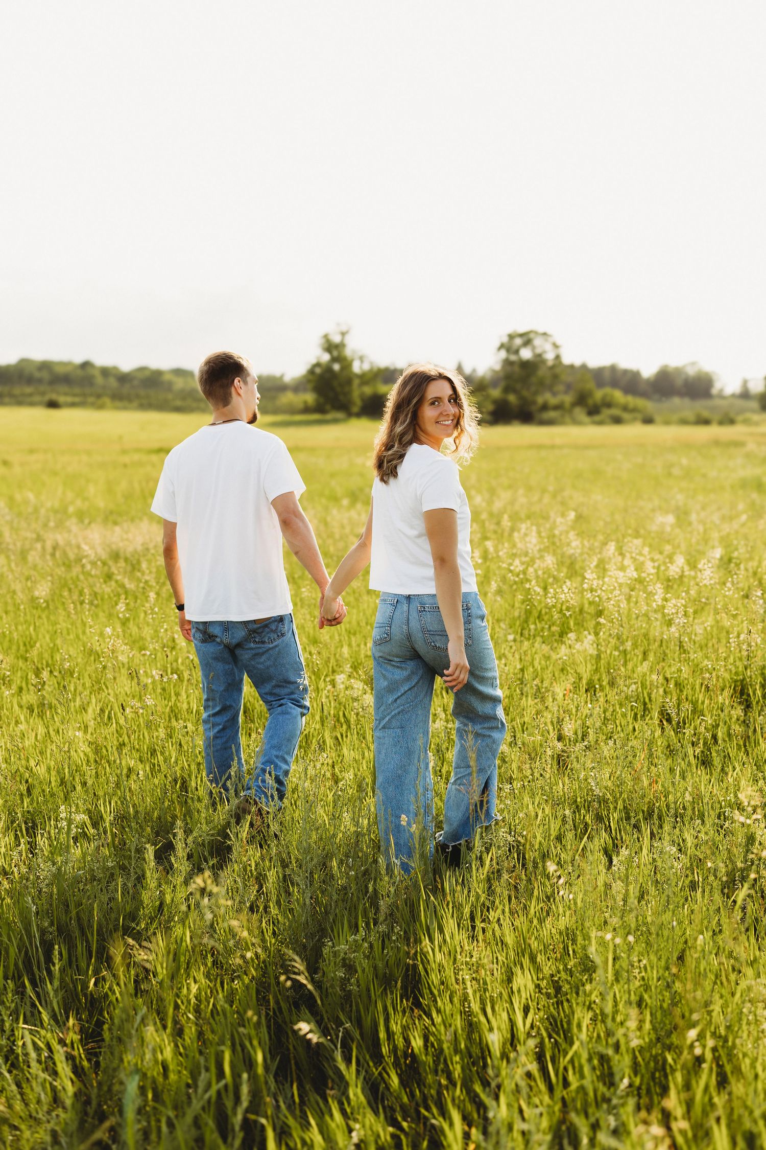 Couple in matching white tees and jeans holding hands in a sunny green field, woman smiling back at camera.