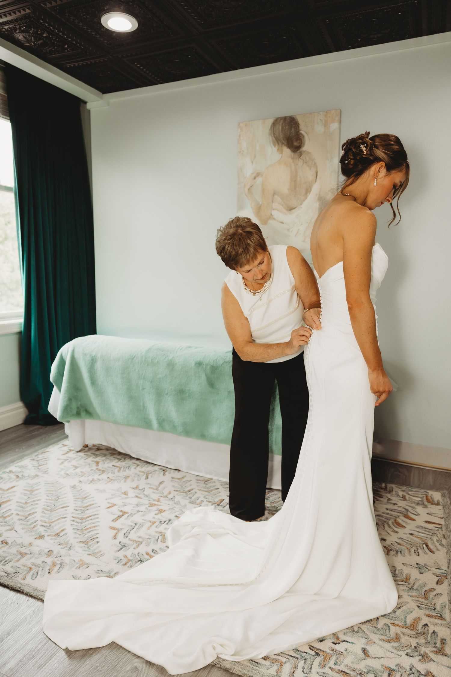 A bride in a white gown gets help fastening her dress from an older woman in a bright hotel suite.