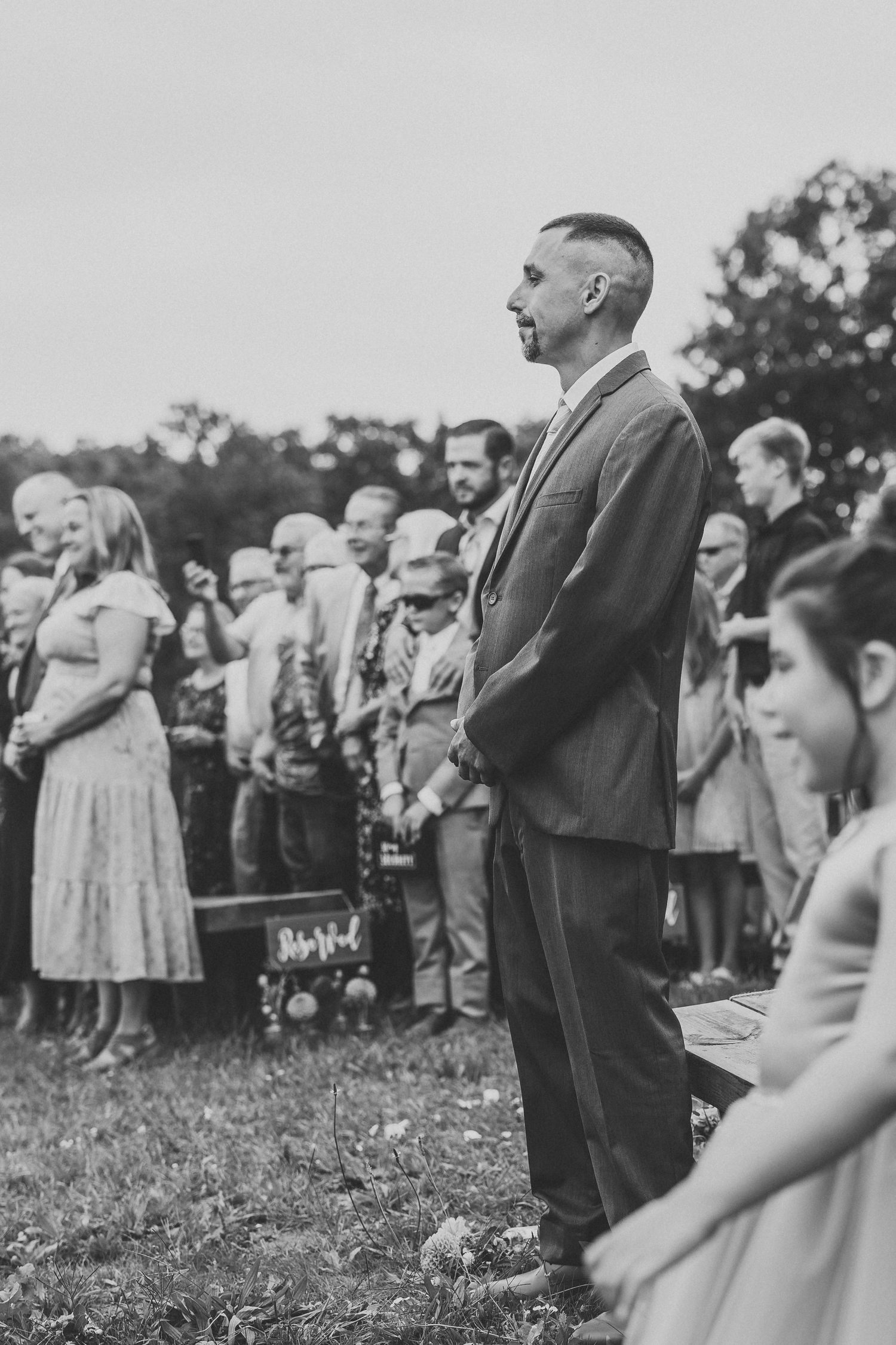 Wedding guests gather outdoors in formal attire for an outdoor ceremony on a grassy field.