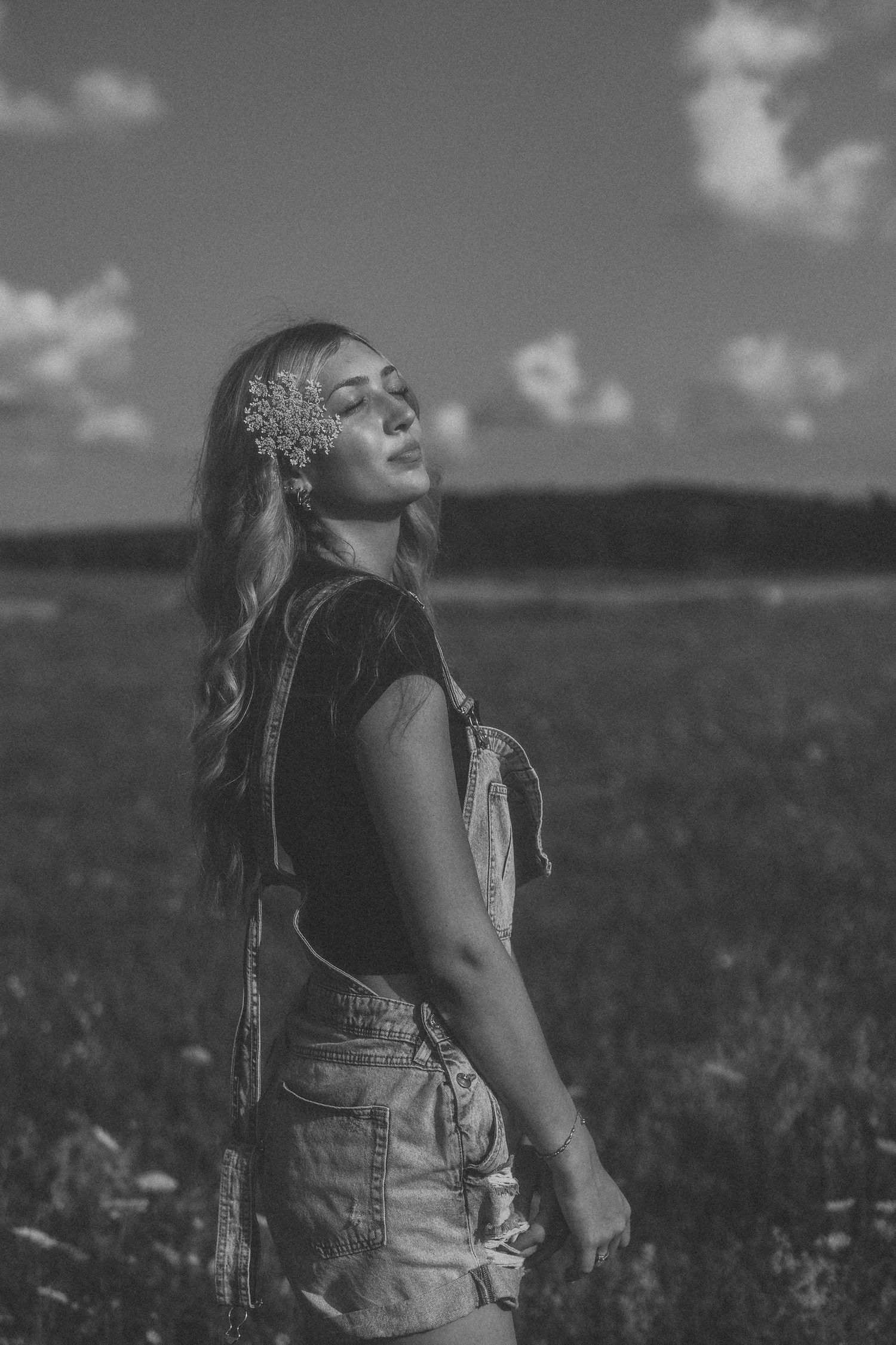 Black and white portrait of someone in overalls with their face turned skyward in a meadow.