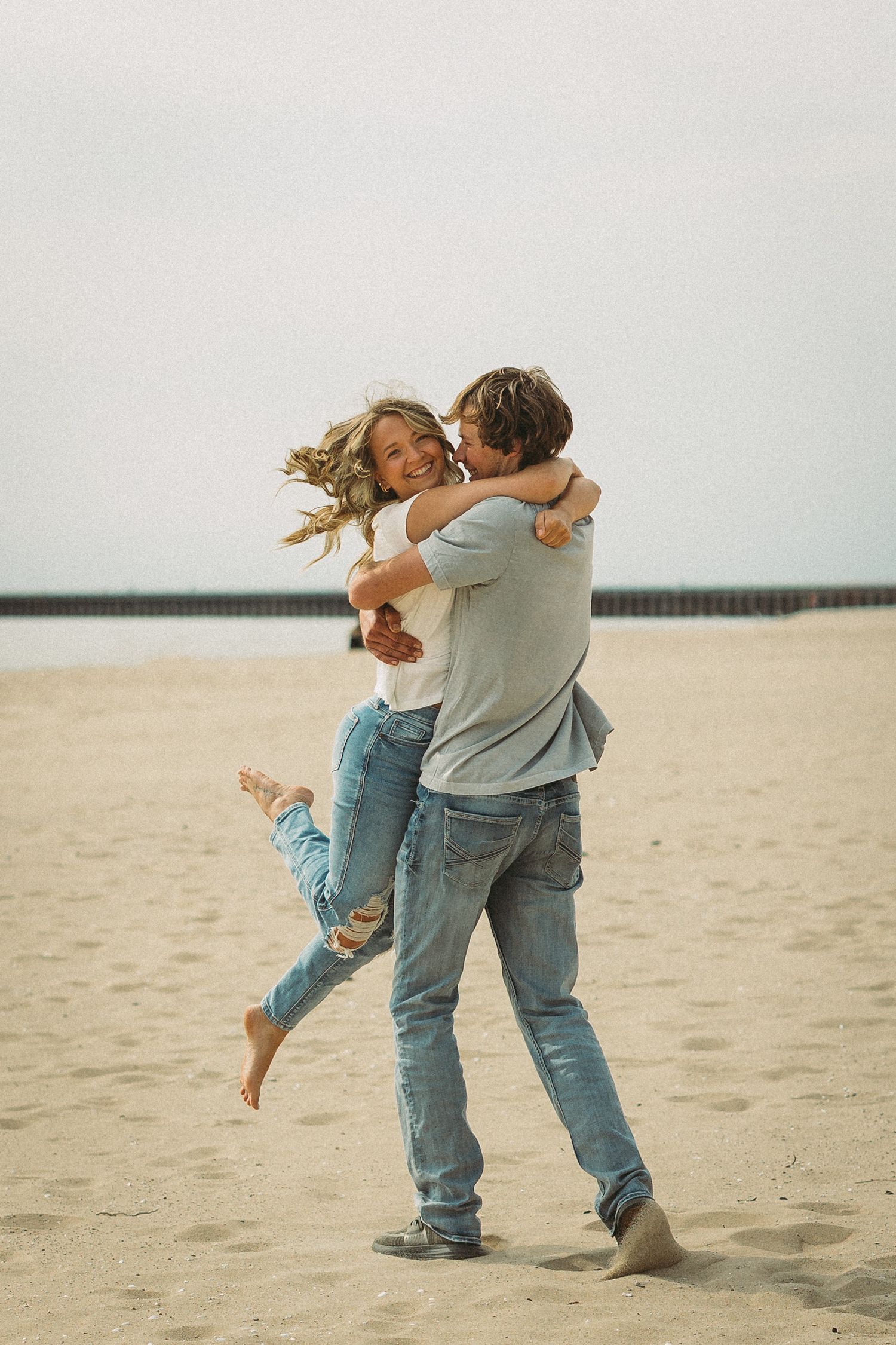 Couple in casual denim sharing a playful dance moment together on a sandy beach.