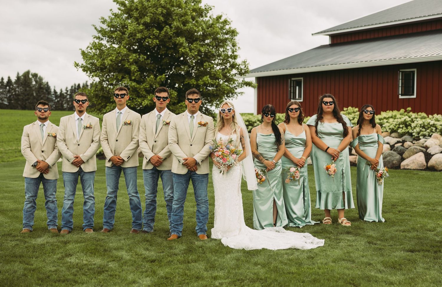 Wedding party poses on grassy lawn with rustic red barn in background during summer celebration.