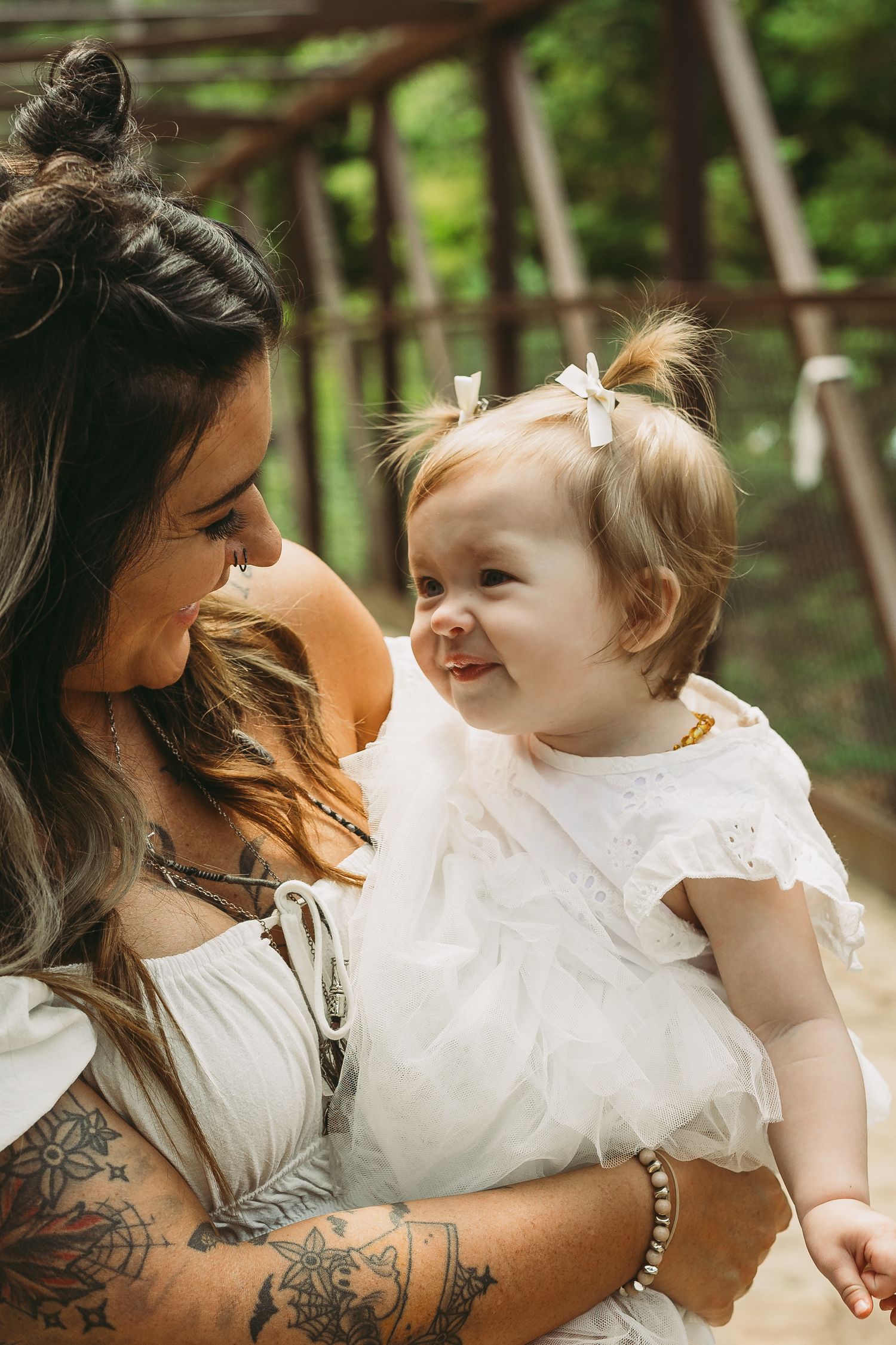 A woman with tattoos and long brown hair holds a baby wearing a white dress in a forest setting.
