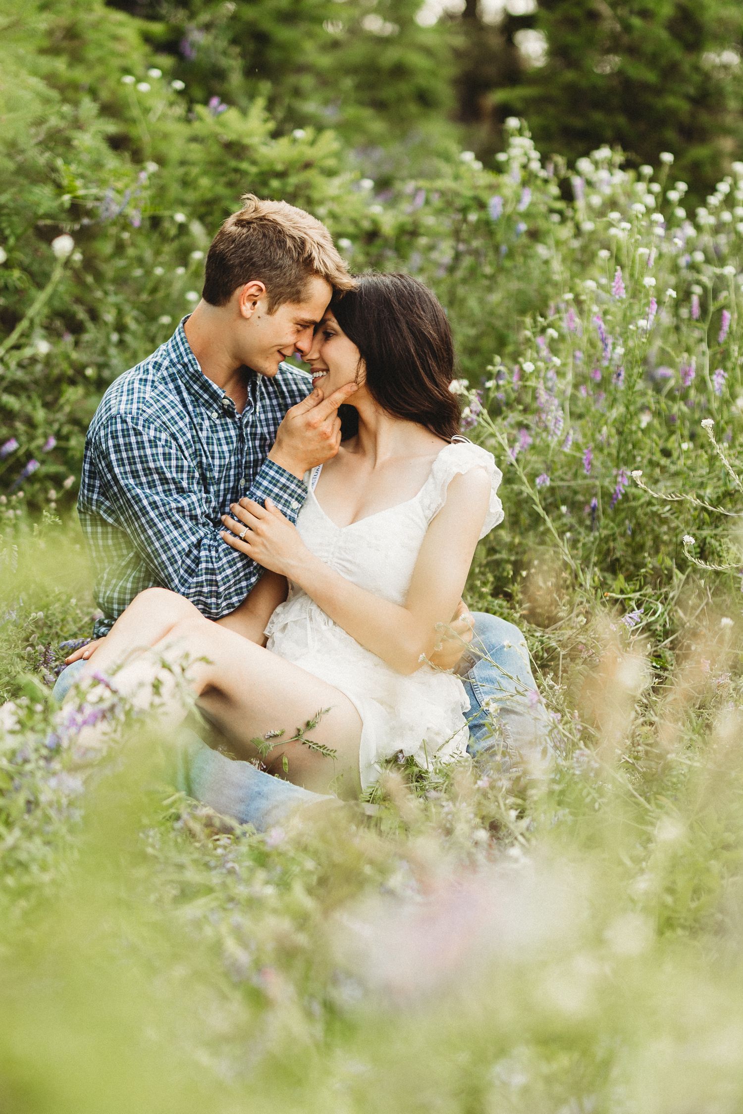 A romantic couple sits intimately among wildflowers in a garden setting during a sunny outdoor photo session.