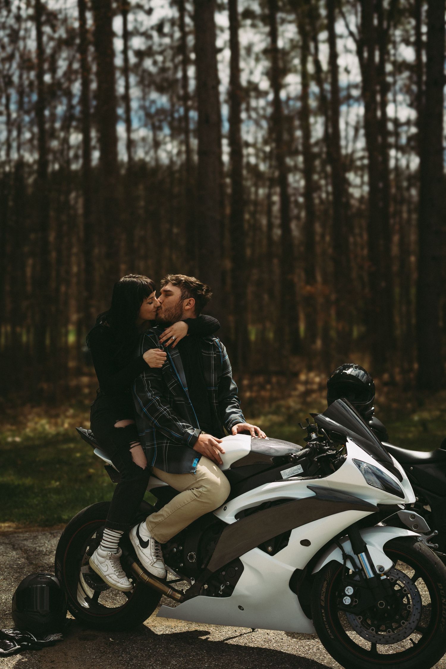 Couple sharing a romantic kiss on a white sport motorcycle parked on a road surrounded by tall pine trees.