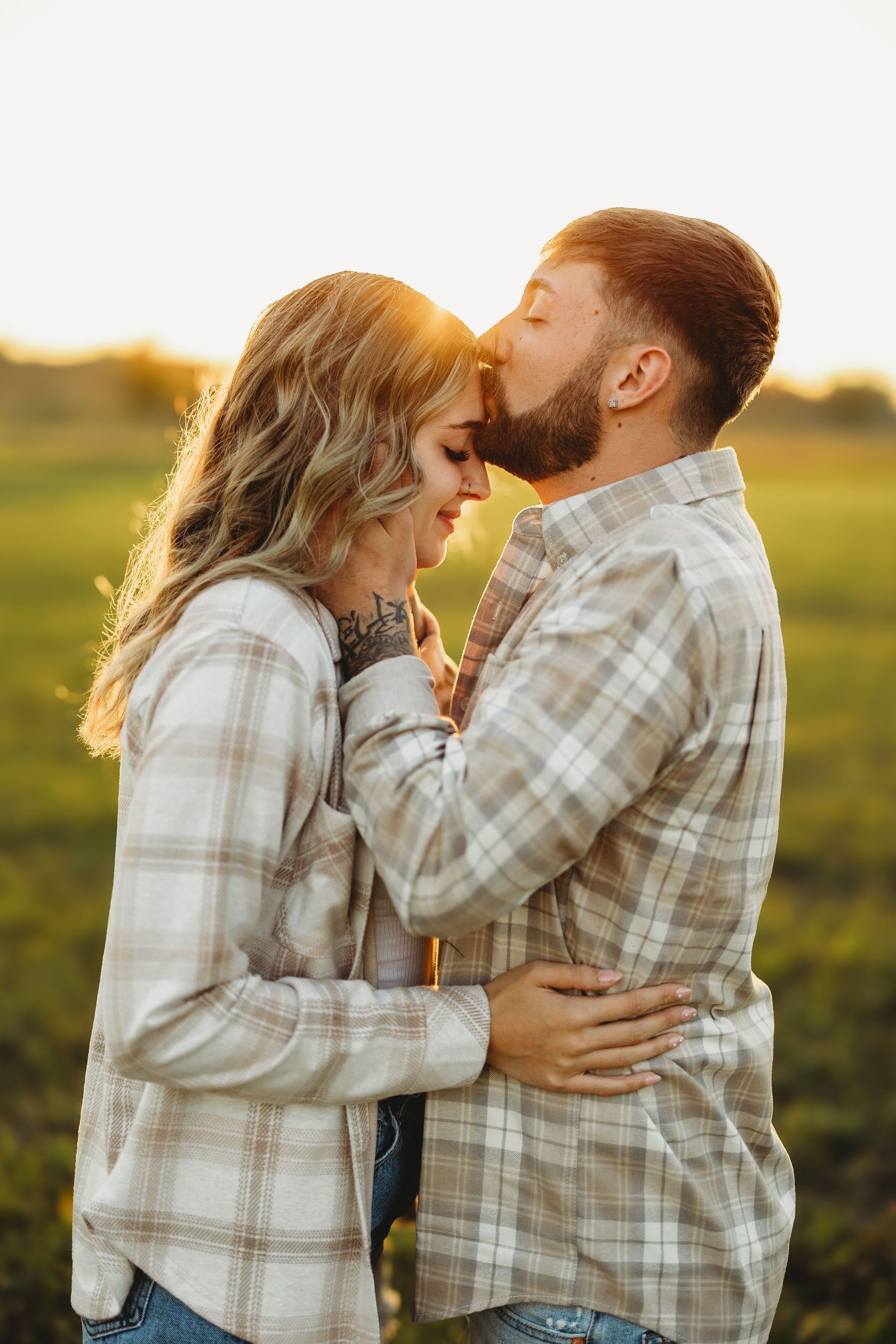 Couple sharing a romantic kiss at golden hour in a green field, both wearing matching plaid shirts.
